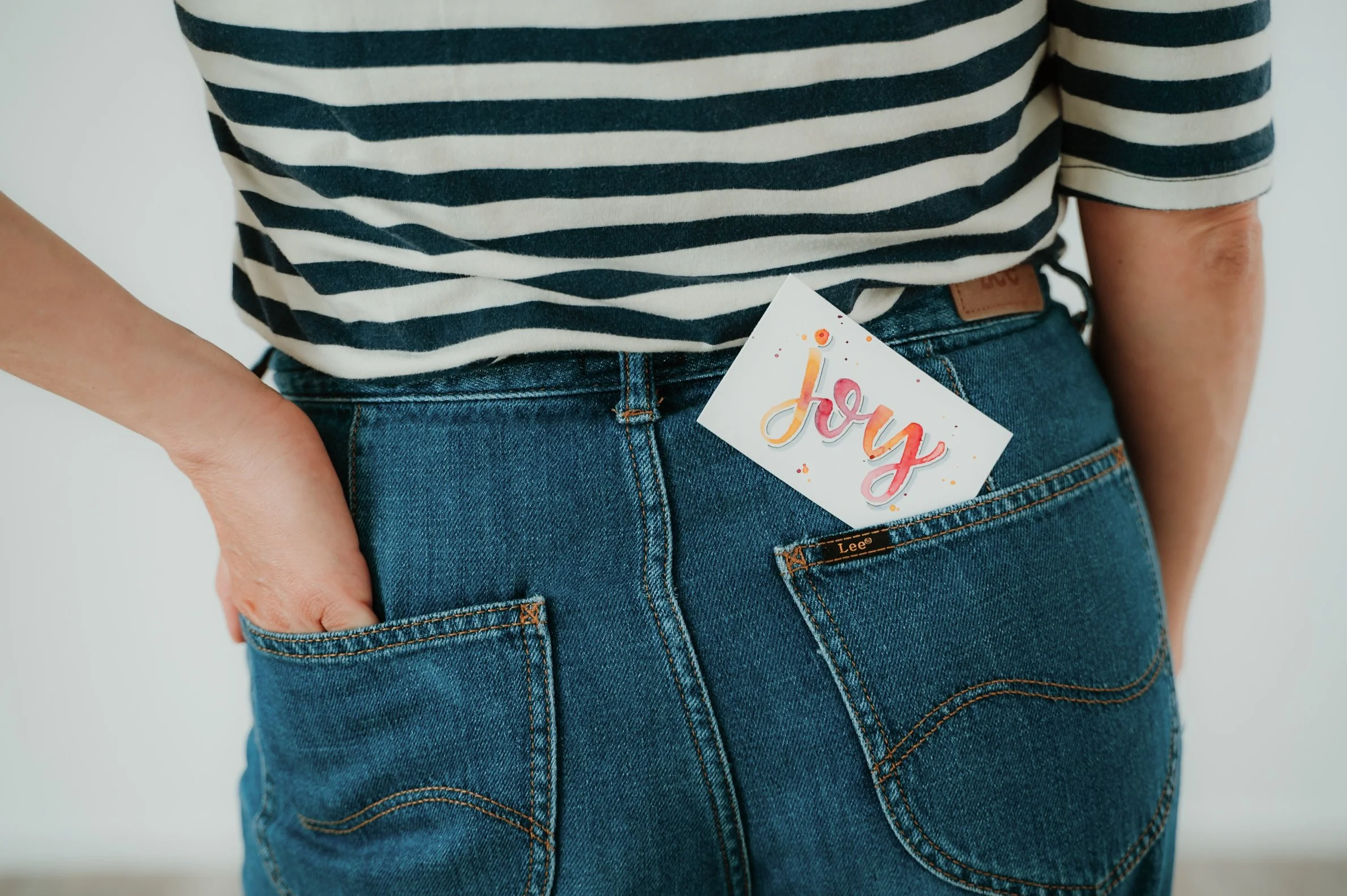 Close-up of a person wearing a black and white striped t-shirt tucked into blue jeans, with a colorful 'joy' sign hanging from the waistband.