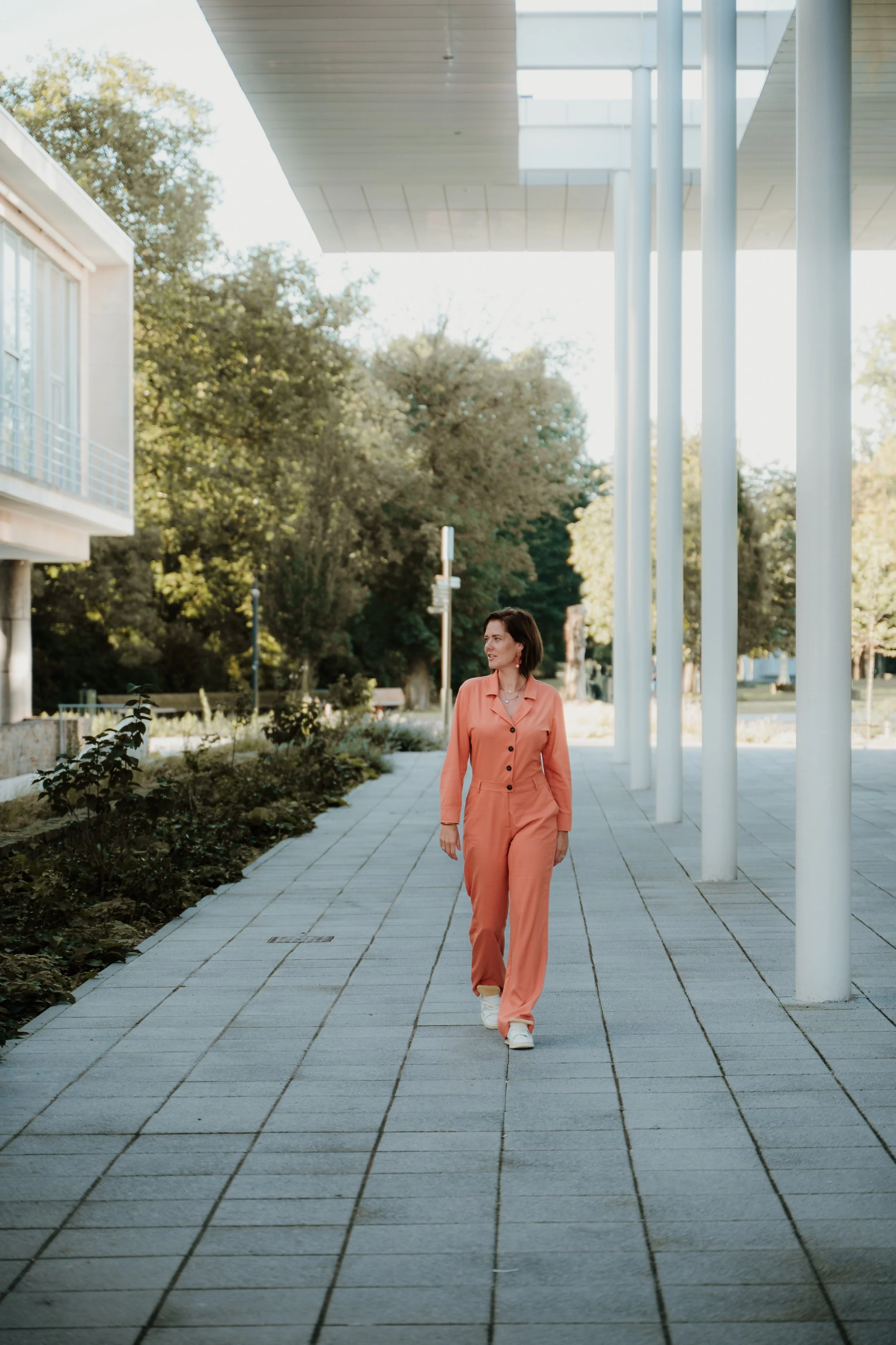 A woman with dark hair walking on a paved sidewalk under a modern building with tall white columns and a roof overhead. She is wearing a peach-colored jumpsuit and white sneakers, with trees and greenery in the background.