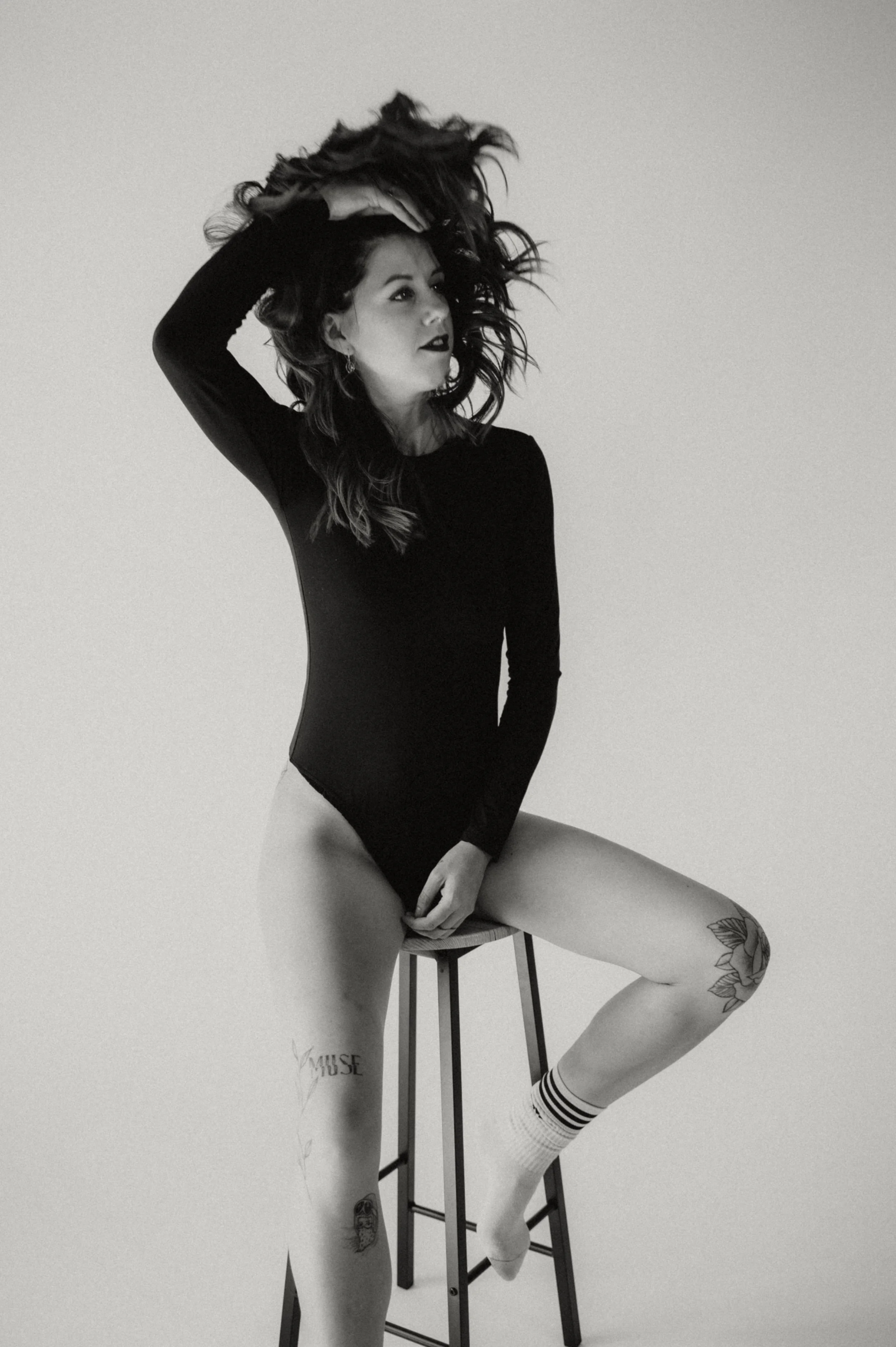 A woman sitting on a stool with one leg raised and her hand on her head, wearing a long-sleeve top, white socks, and underwear, with tattoos visible on her thigh and arm, in a black-and-white photo against a plain background.
