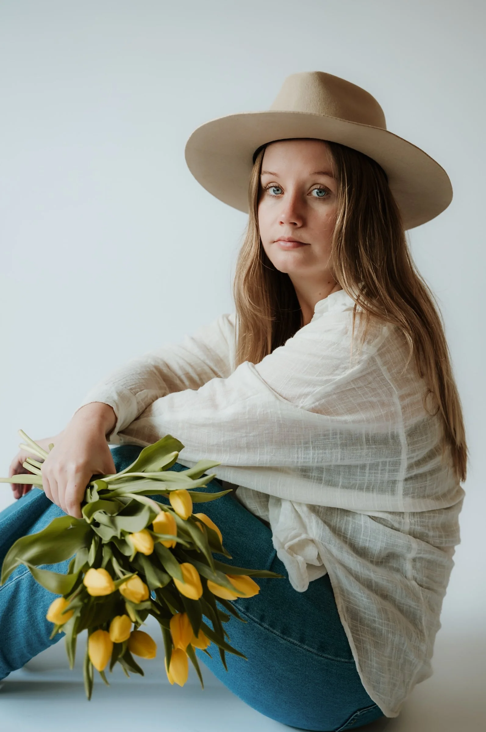 A woman wearing a beige wide-brimmed hat and a light-colored blouse sitting and holding a bouquet of yellow tulips. She is seated on the floor with a neutral background.