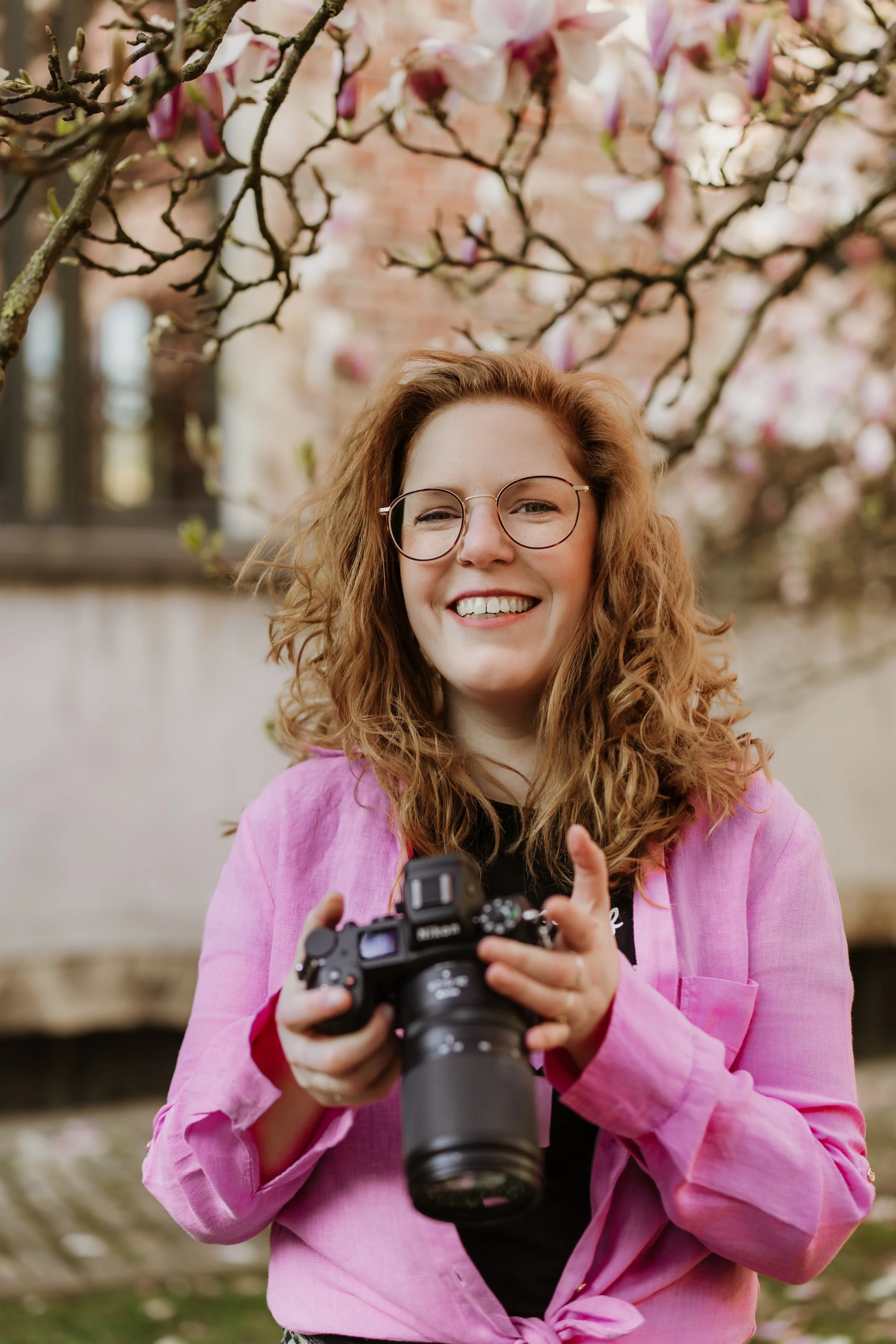 A young woman with curly red hair and glasses smiling while holding a camera, standing outdoors in front of blooming pink and white magnolia trees.