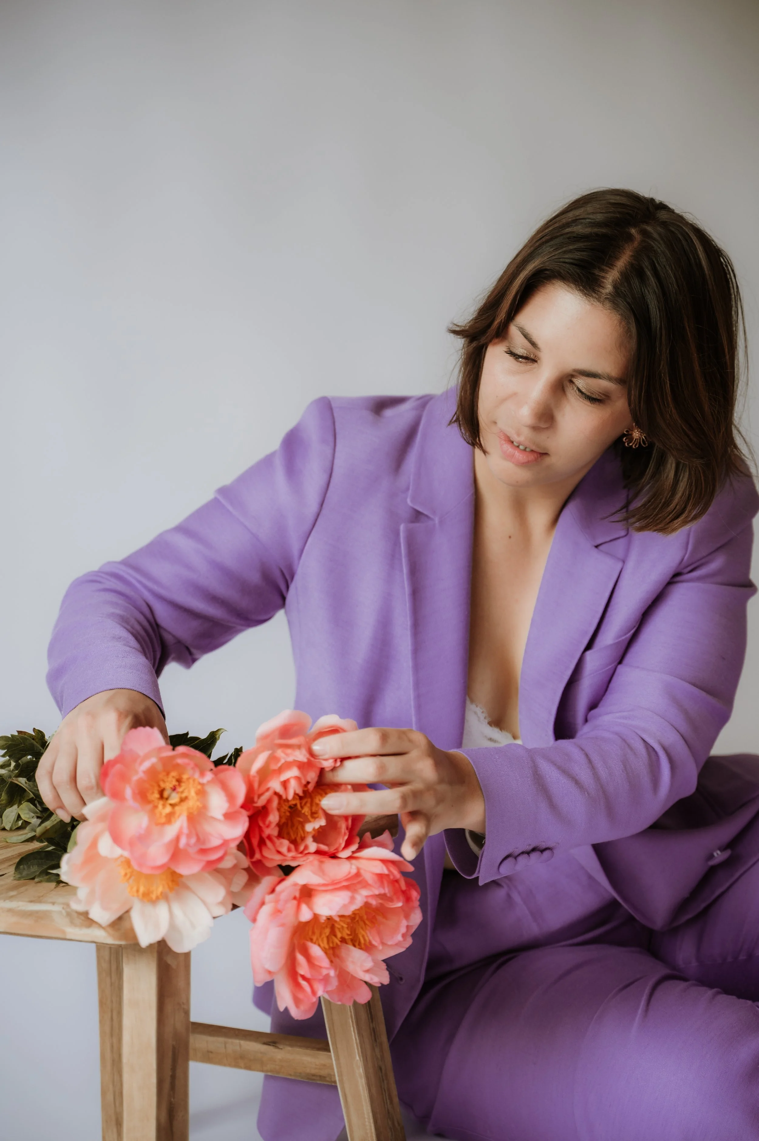 A woman in a purple blazer arranges pink and orange peony flowers on a wooden table.