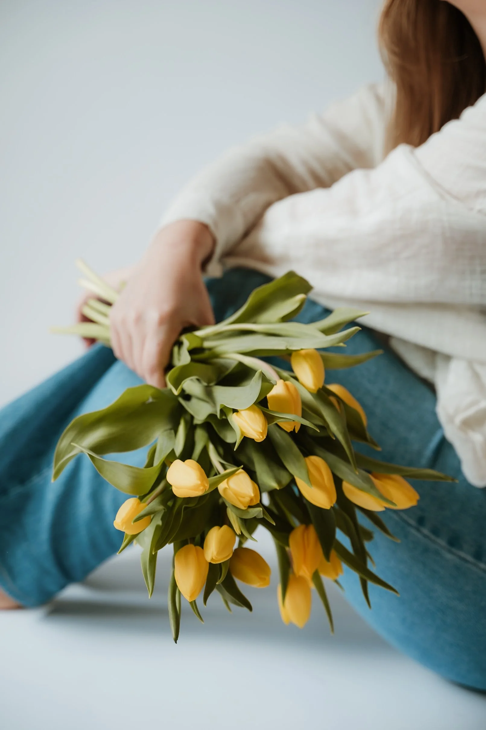 Person sitting with crossed arms holding a bouquet of yellow tulips, wearing a beige long-sleeve shirt and blue jeans, against a plain light background.