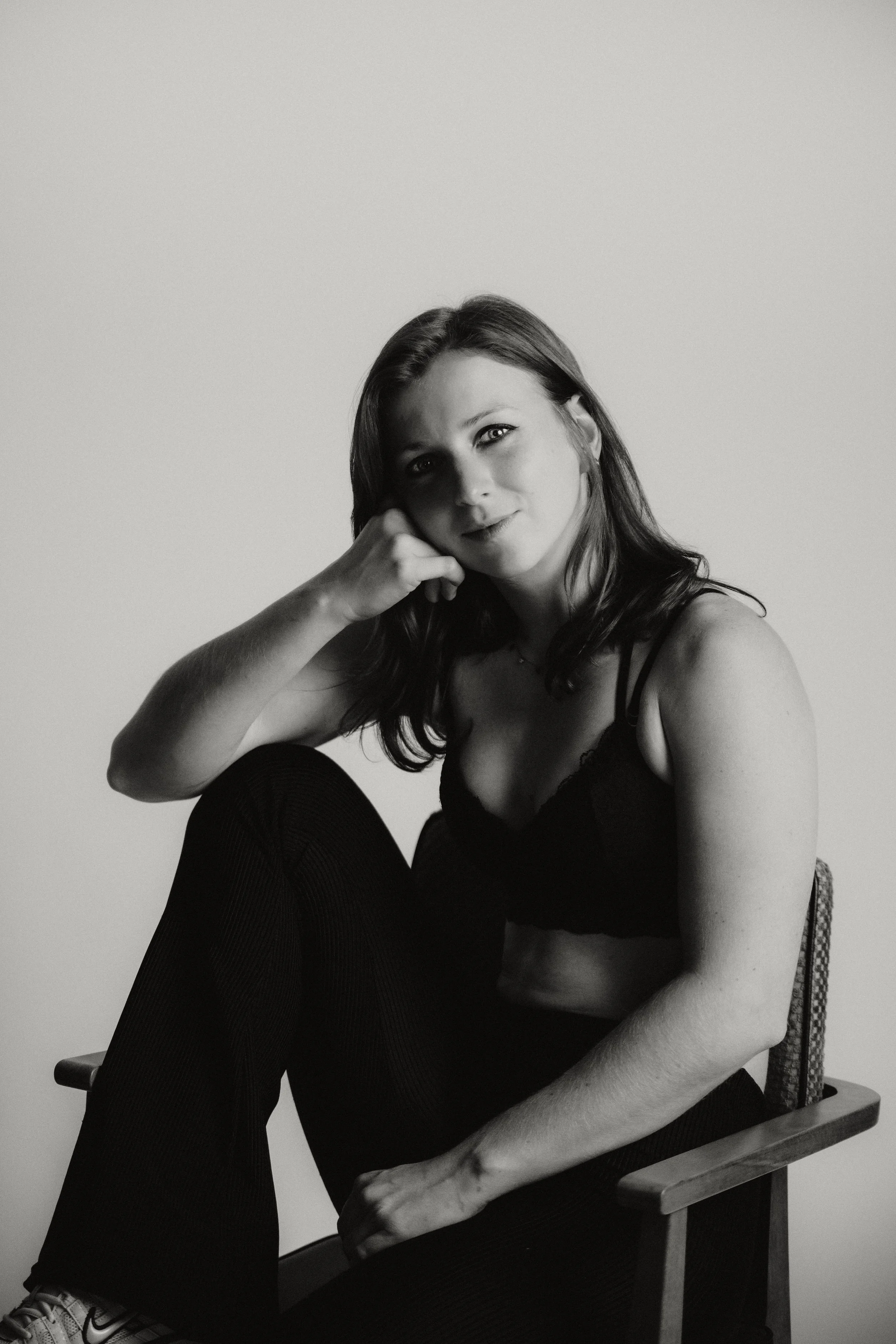 Black and white photo of a young woman with dark hair, sitting on a chair, wearing a black crop top and leggings, posing with her elbow resting on her knee, looking at the camera.