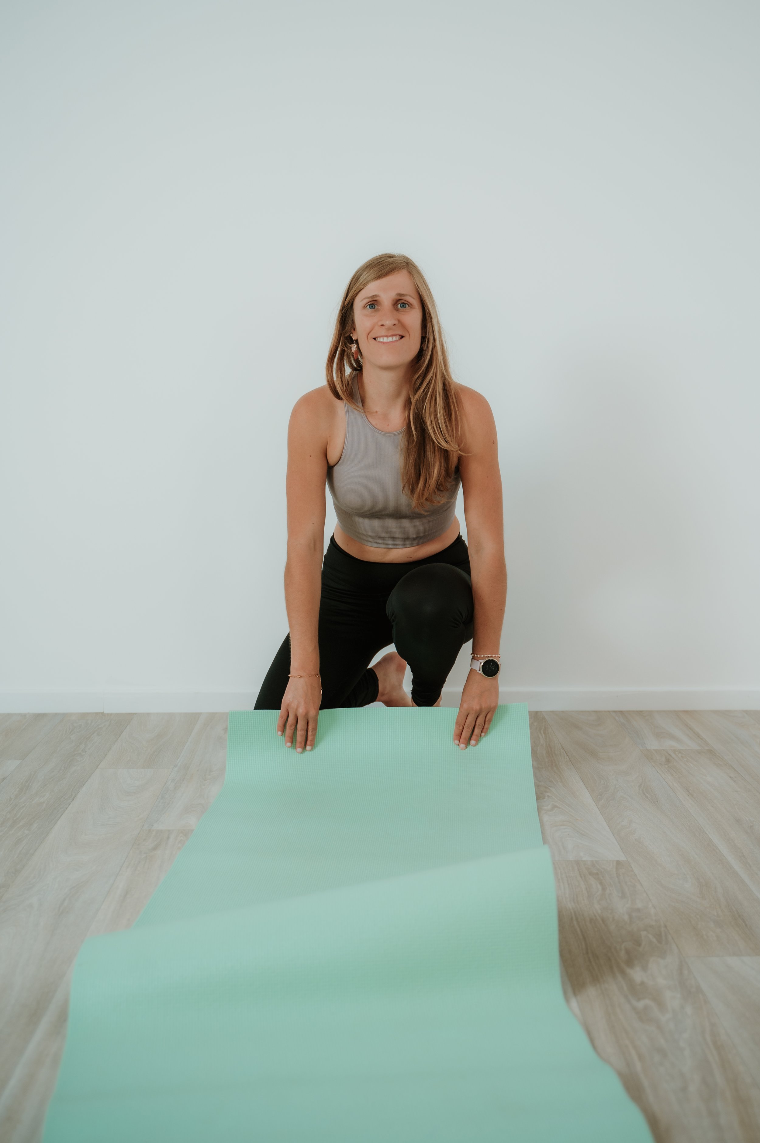 A woman practicing yoga or stretching on a light green yoga mat indoors, kneeling with one knee on the floor and the other foot flat on the ground, wearing a sleeveless gray top, black pants, a watch on her left wrist, and an earring, with a plain wh