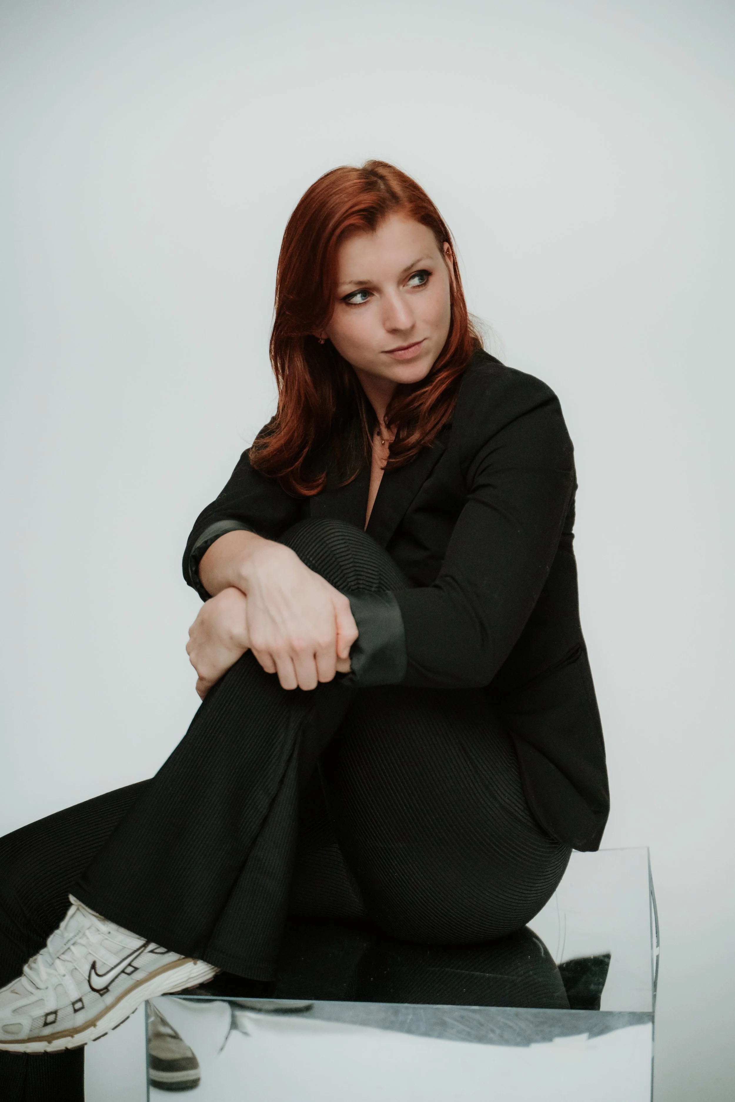 A young woman with red hair and light skin sitting on a reflective surface or mirror, wearing a black blazer, black pants with a subtle striped pattern, and white sneakers with black accents. She looks to her left with a neutral expression against a 