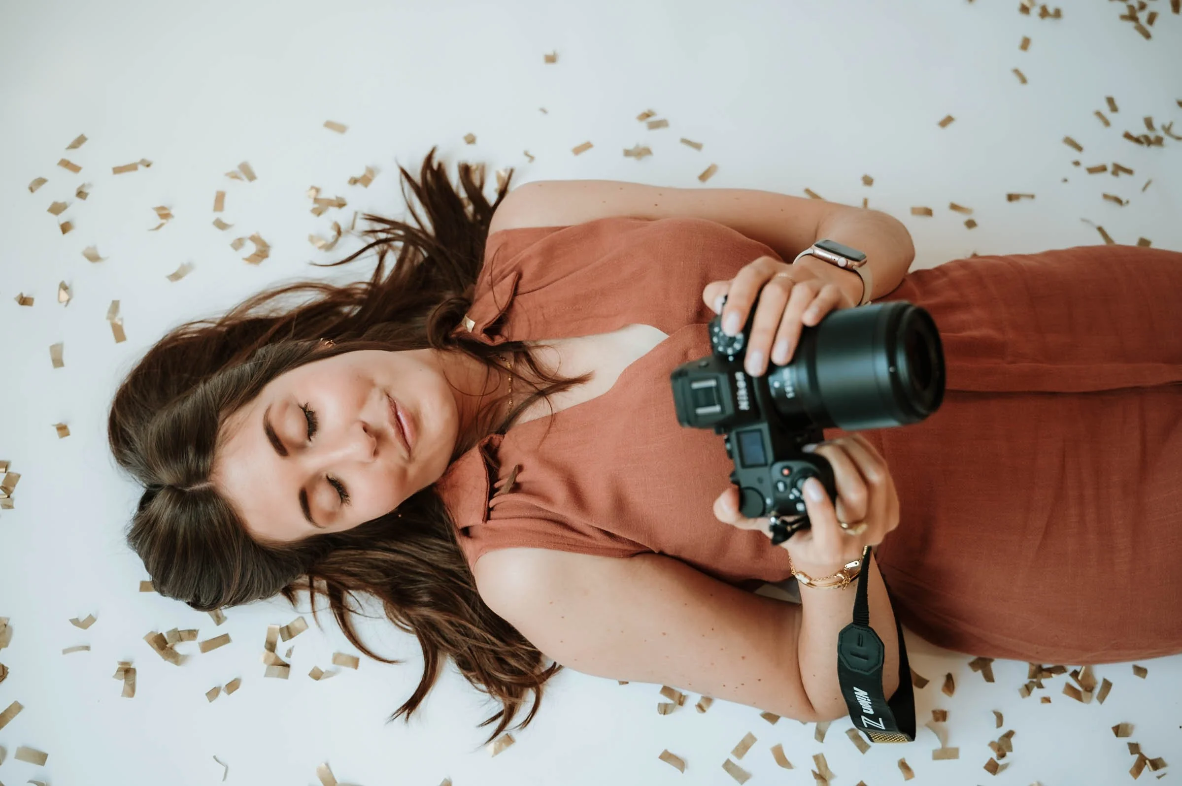 A woman with long brown hair lying down covered in confetti, holding a camera with her eyes closed, wearing a rust-colored dress and a smartwatch.