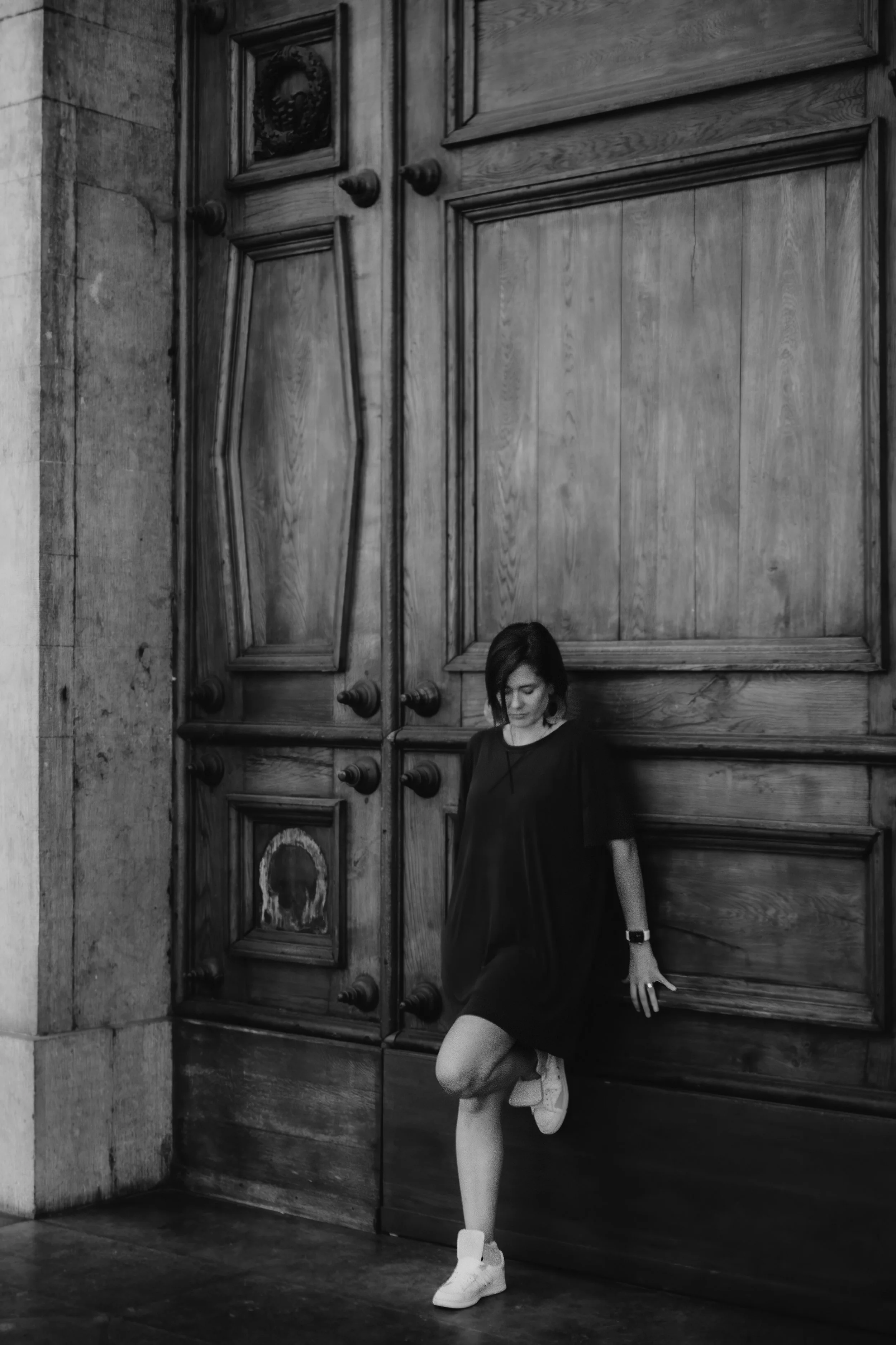 A young woman with short dark hair wearing a black dress, white sneakers, fishnet stockings, and a watch, standing against a large wooden door with ornate carvings and carved knobs, in an indoor setting.