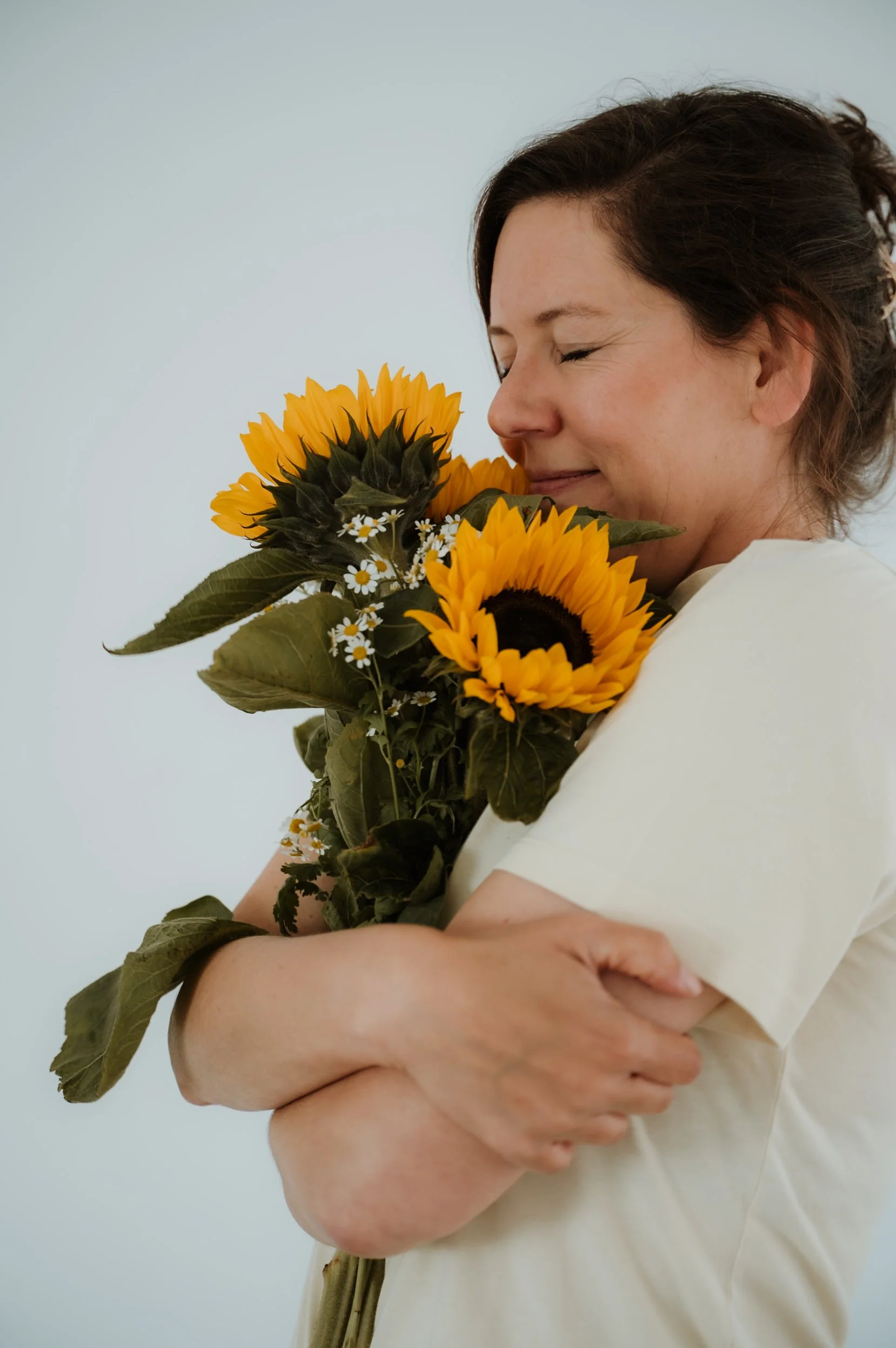A woman with brown hair and closed eyes holds a bouquet of sunflowers and small white daisies, standing against a plain light blue background.