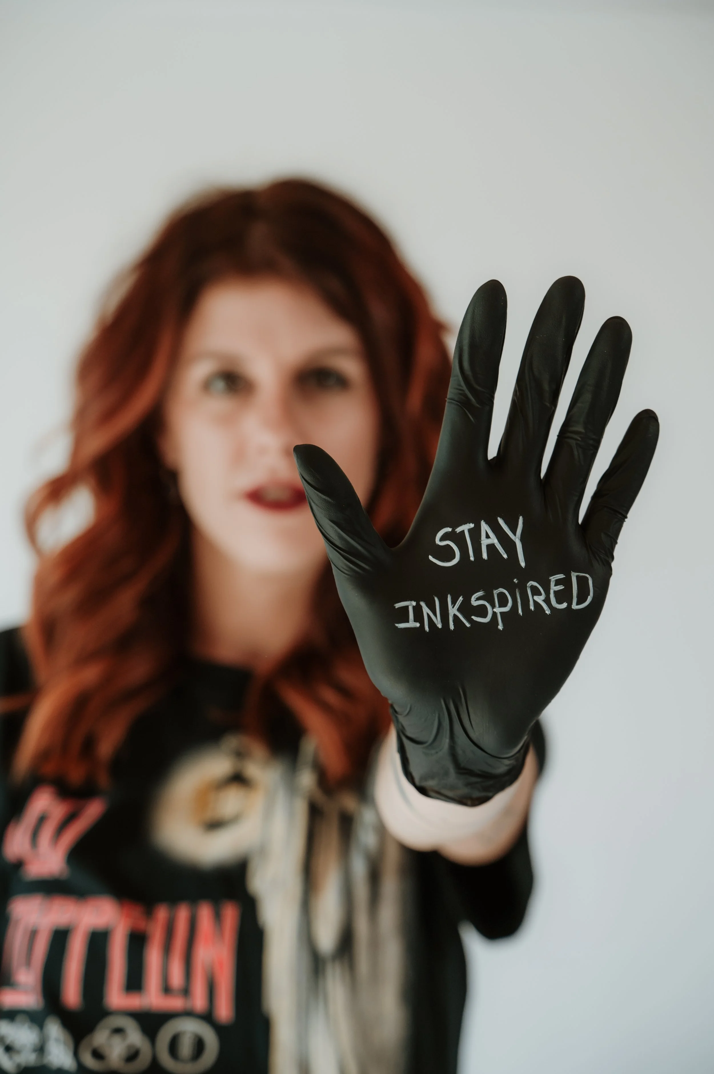 A woman with red hair holding up a black glove with the words 'STAY INSPIRED' written on it, in front of a blurred background.