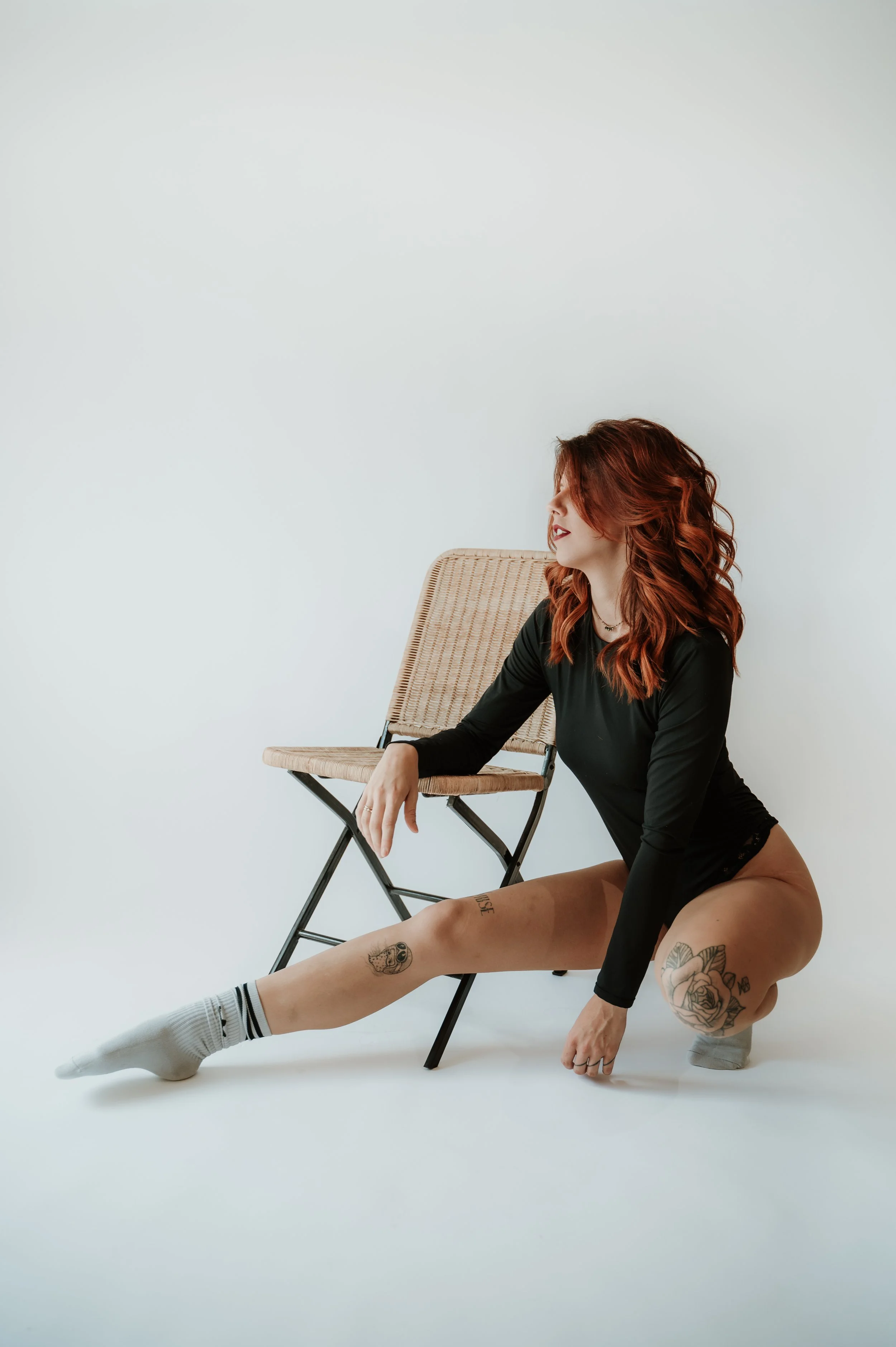 A woman with red wavy hair and tattoos on her legs is sitting on the floor with her upper body turned to the side, one arm resting on a chair. She is wearing a black long-sleeve top, gray socks, and is positioned next to a folding chair against a pla