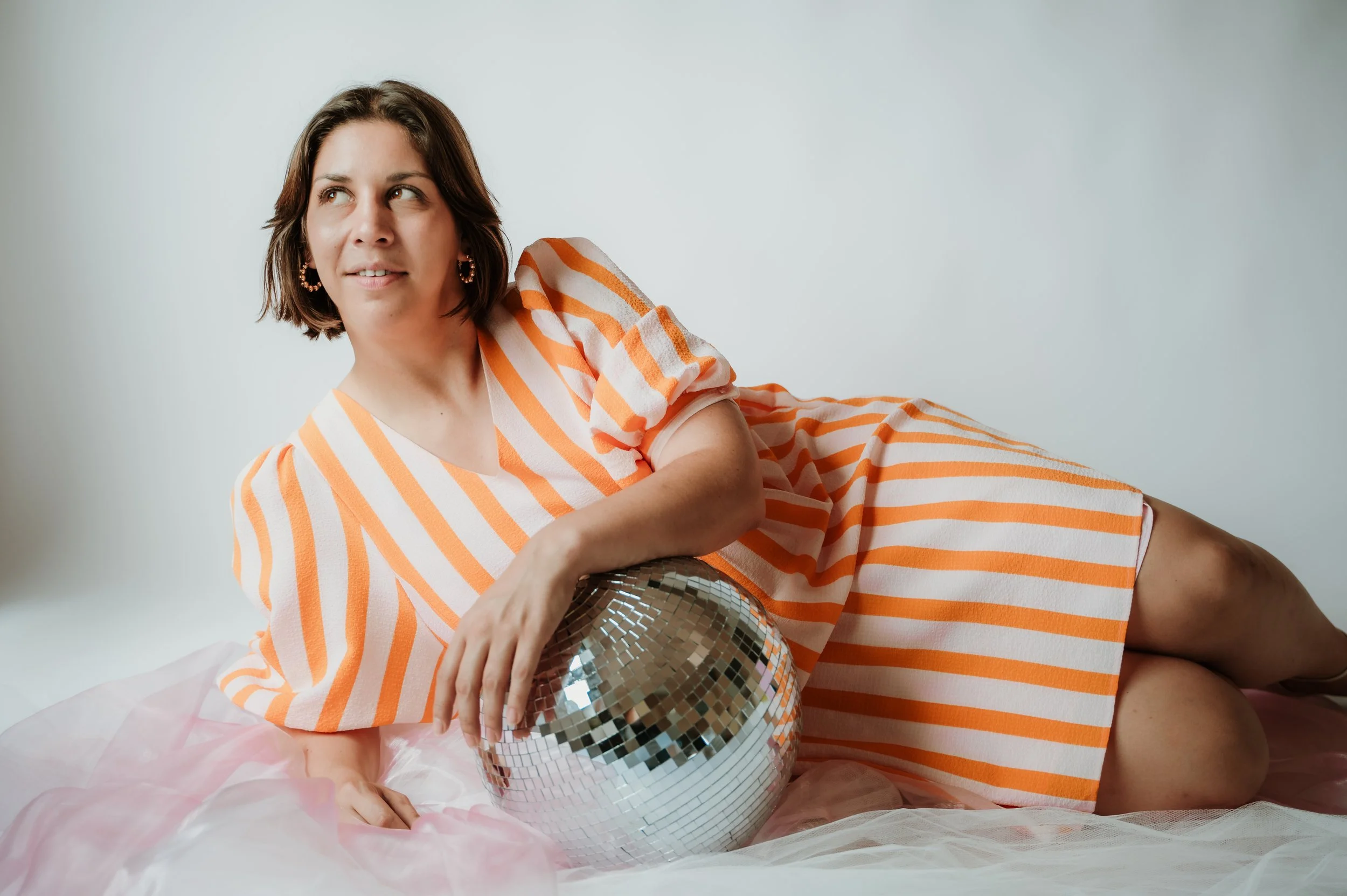 A woman with short brown hair wearing a striped orange and white dress, lying on pink fabric, holding a disco ball and looking to the side against a plain light background.