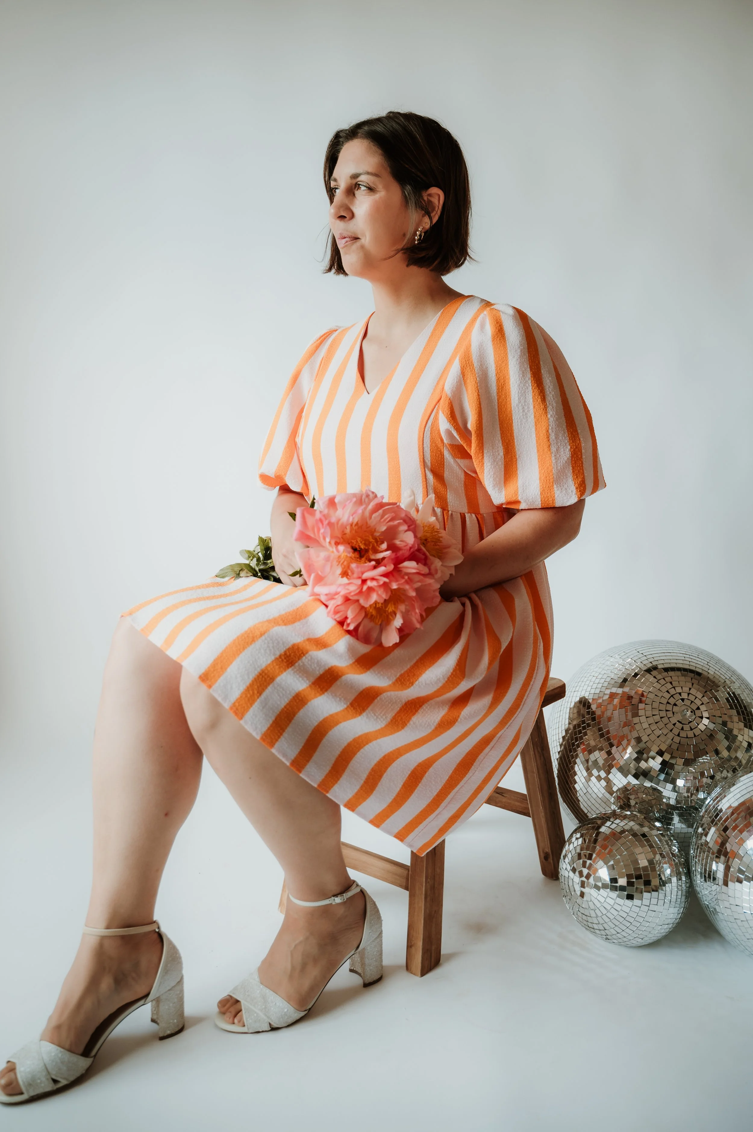 A woman with dark hair and earrings wearing a striped orange and white dress is sitting on a wooden chair holding pink flowers. She is wearing high-heeled shoes and sitting with one leg crossed over the other. Next to her on the floor are several dis
