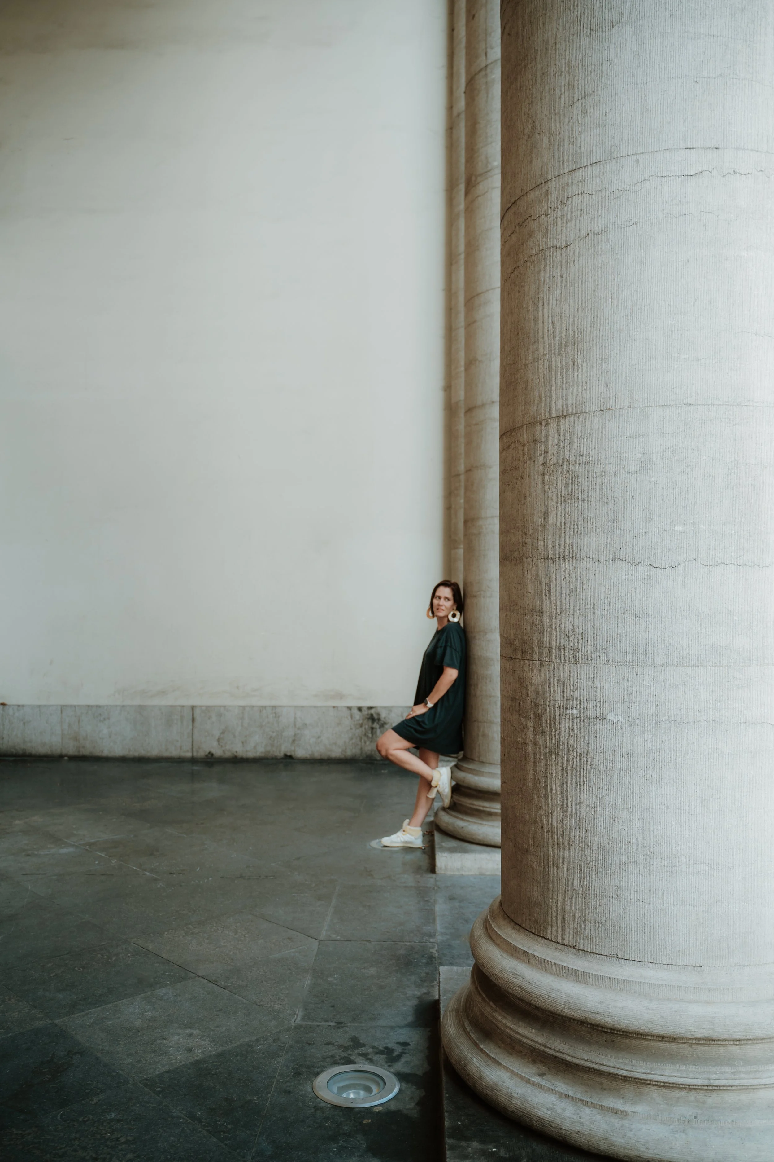 A woman in a dark green dress and white sneakers is standing and leaning against a large round stone column in a minimalistic space with a blank wall behind her and a black floor.