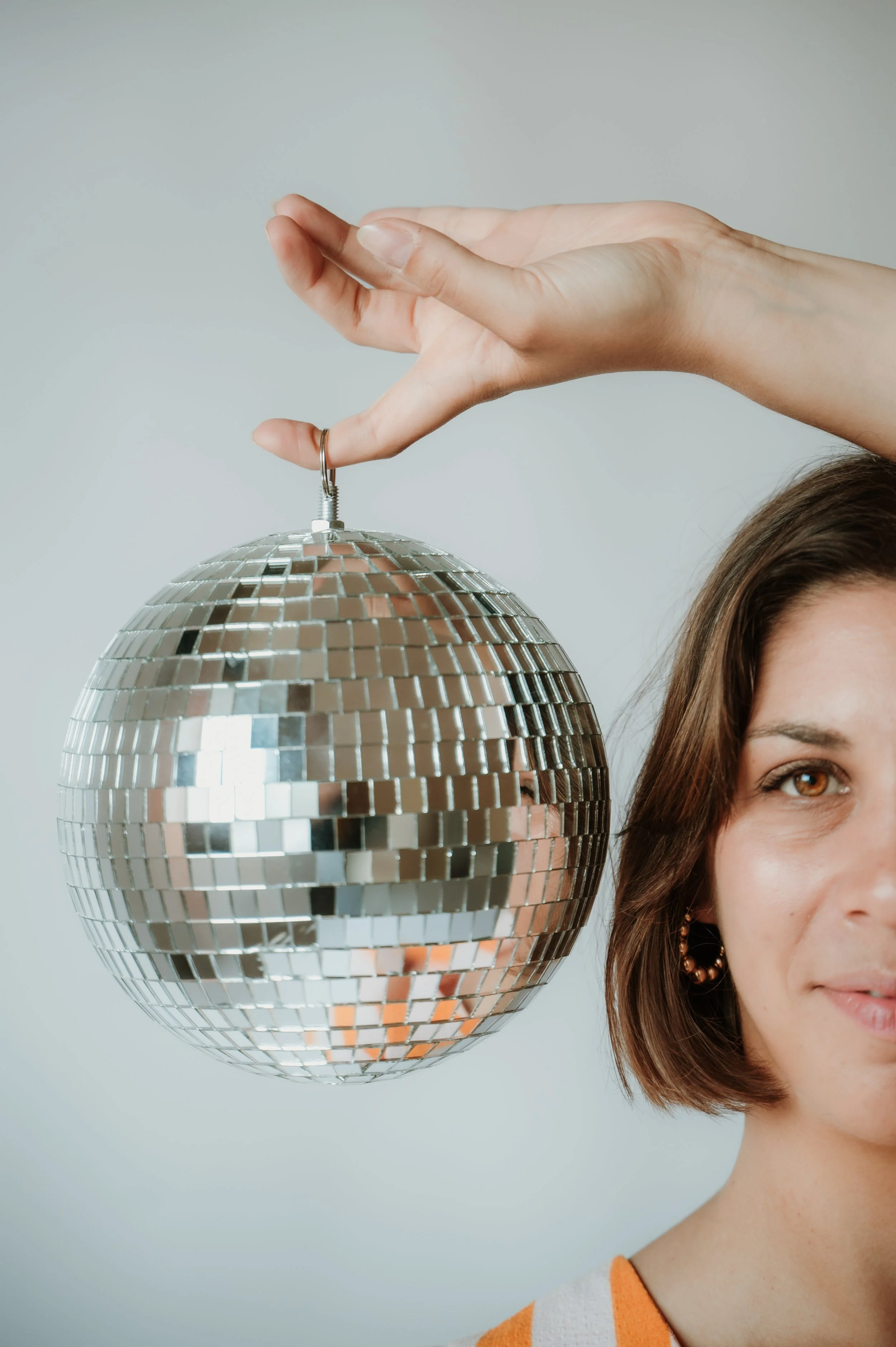 A woman with brown hair and brown eyes wearing a striped orange and white top standing next to a shiny silver disco ball hanging from the ceiling.