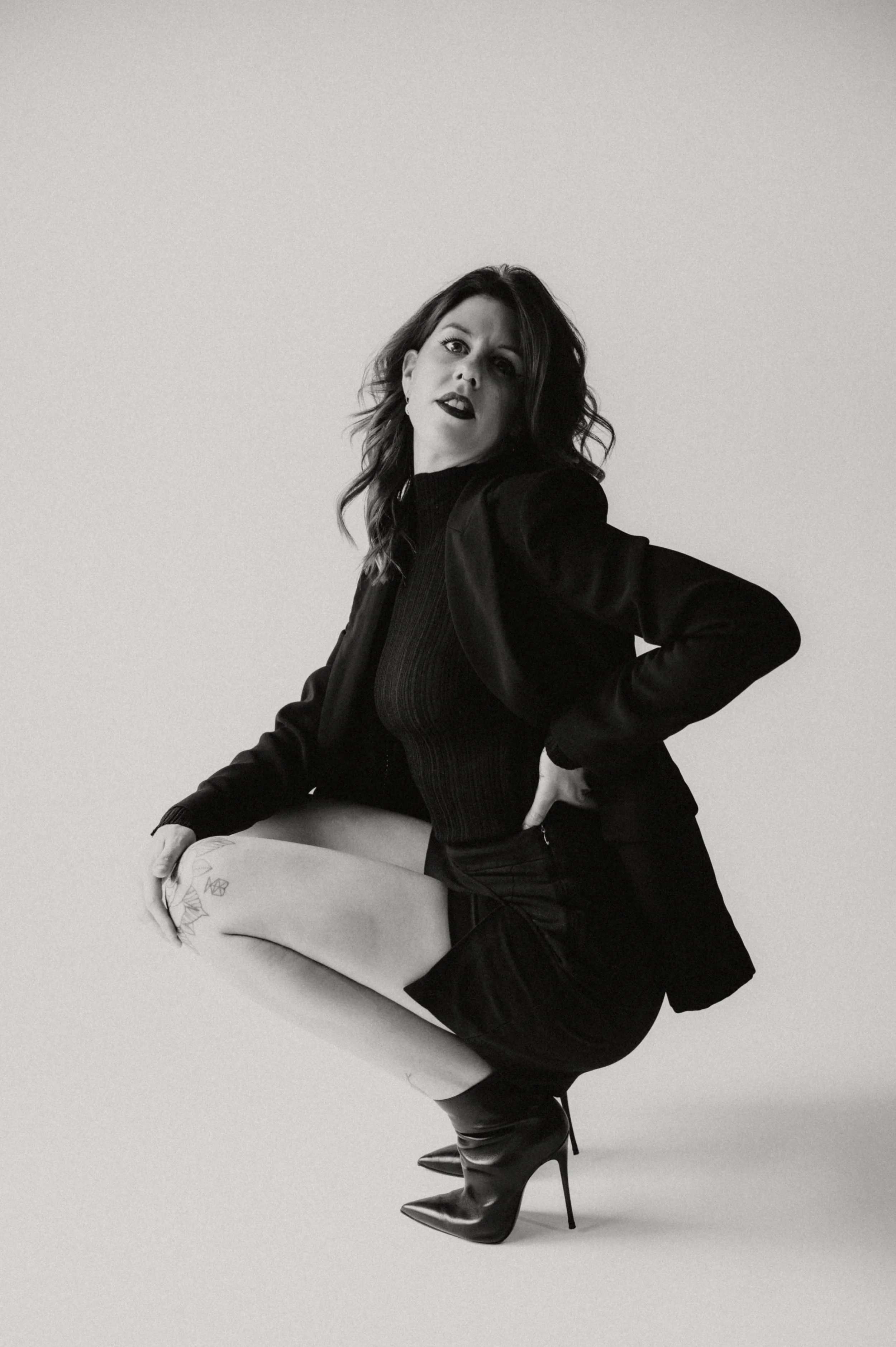 Black and white photo of a woman in high heels, leather jacket, and lipstick, with her left hand on her hip and her right arm resting on her knee, sitting on a concrete surface.