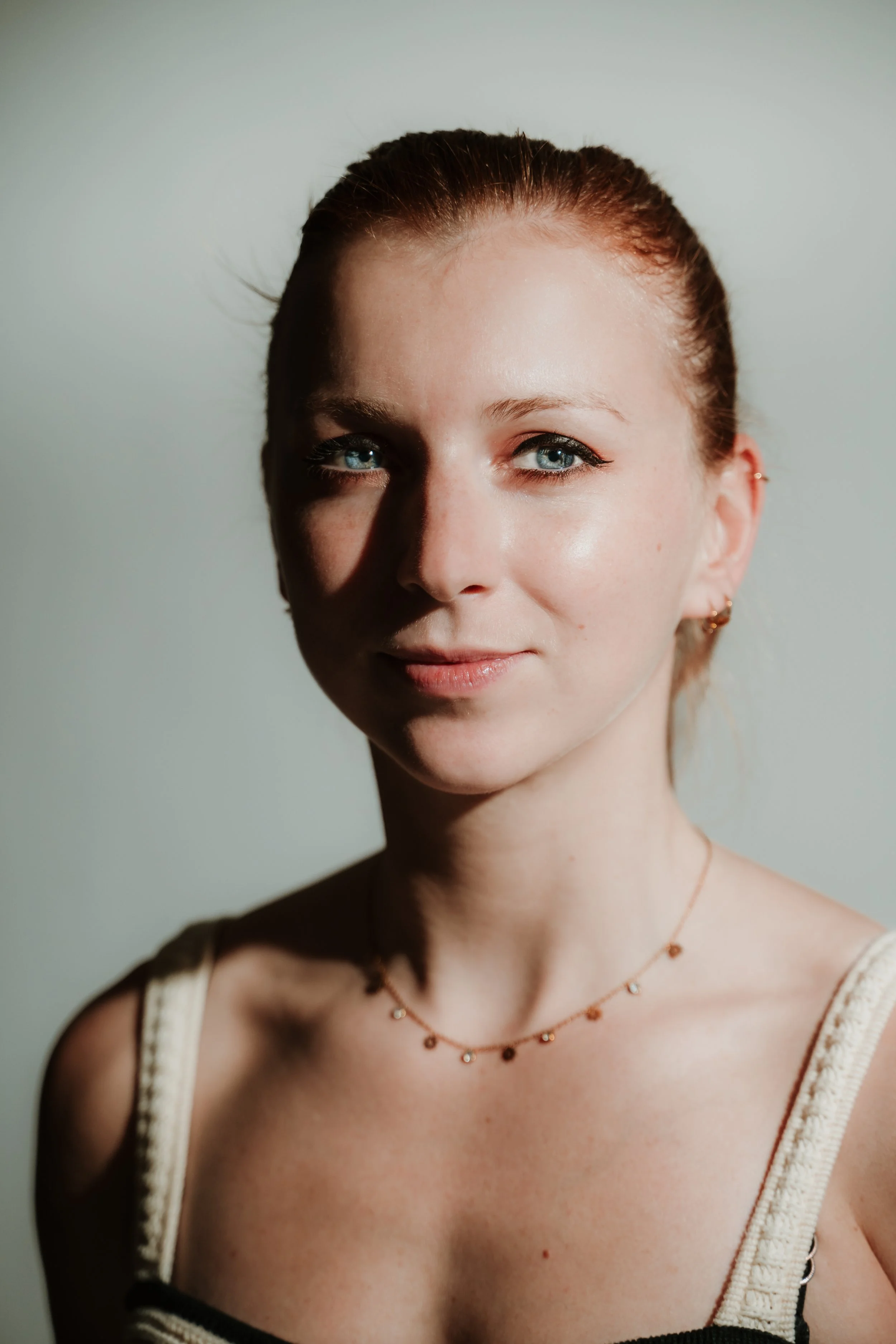 Close-up of a woman's face, showing her blue eyes, red hair tied back, small earrings, a delicate necklace, and wearing a cream-colored top with black straps, against a plain background.