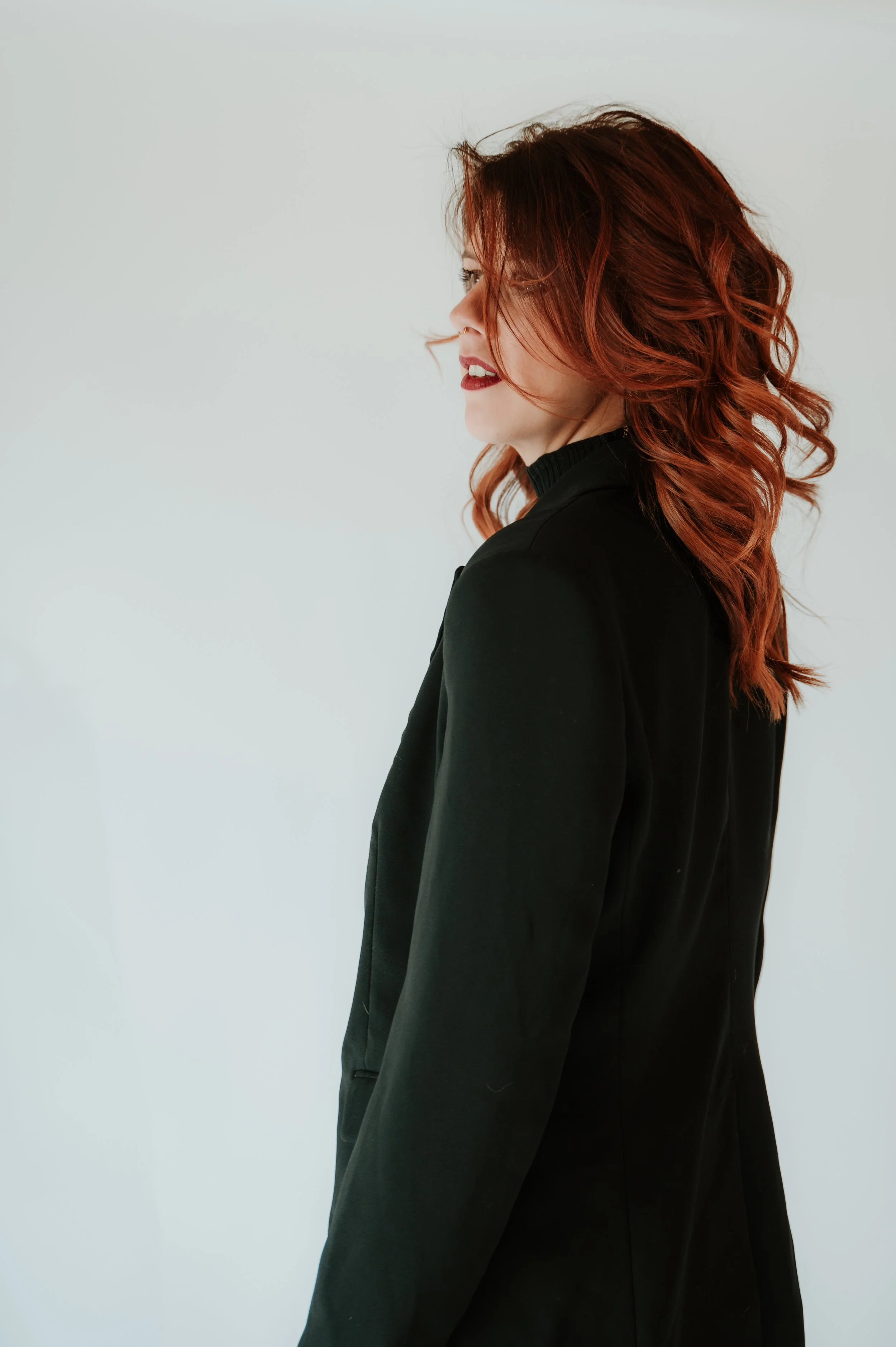 Side view of a woman with long curly red hair wearing a black blazer, standing against a plain light gray background.