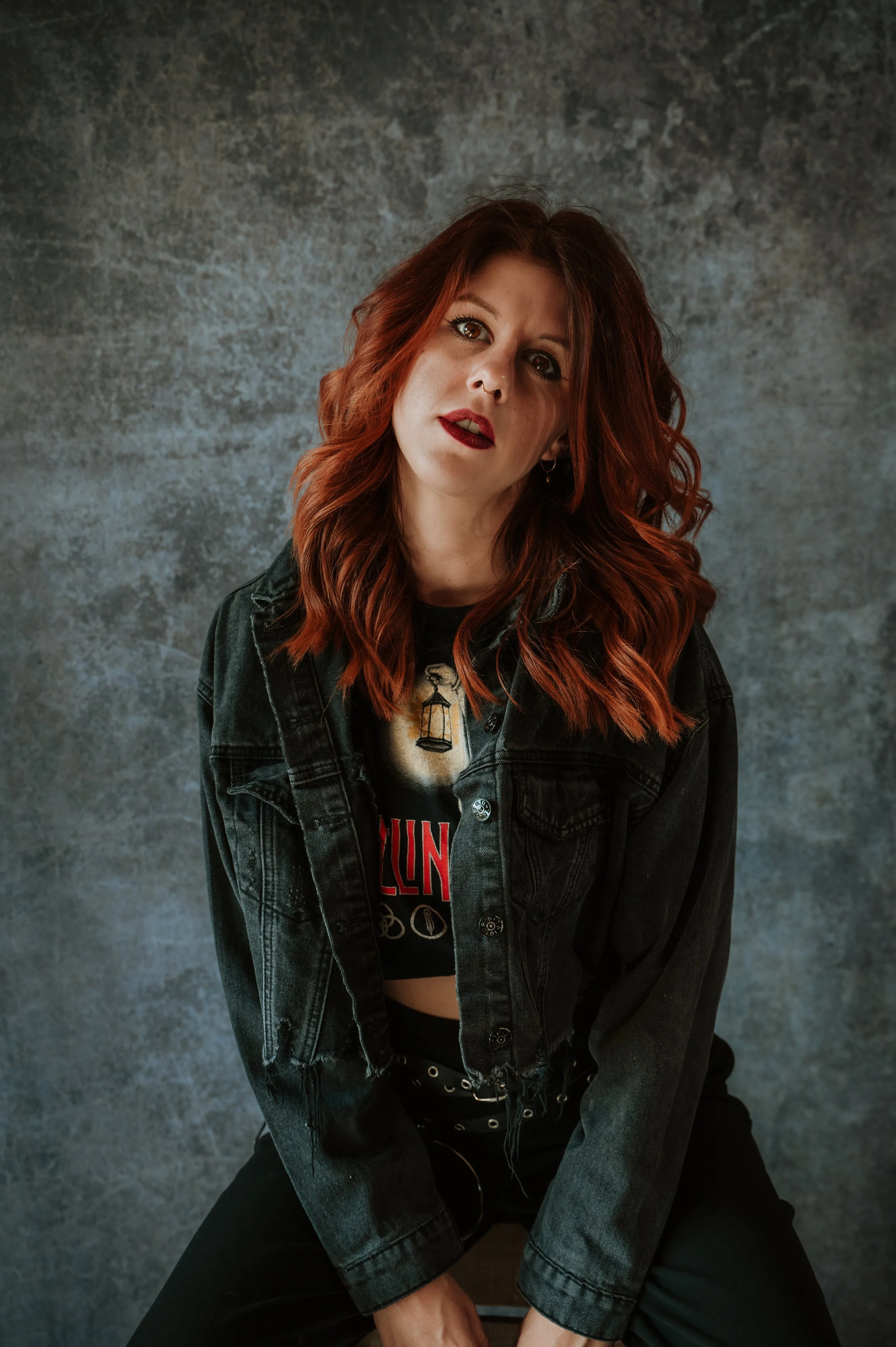 A woman with wavy red hair wearing a black denim jacket and a graphic t-shirt, sitting against a textured gray background.
