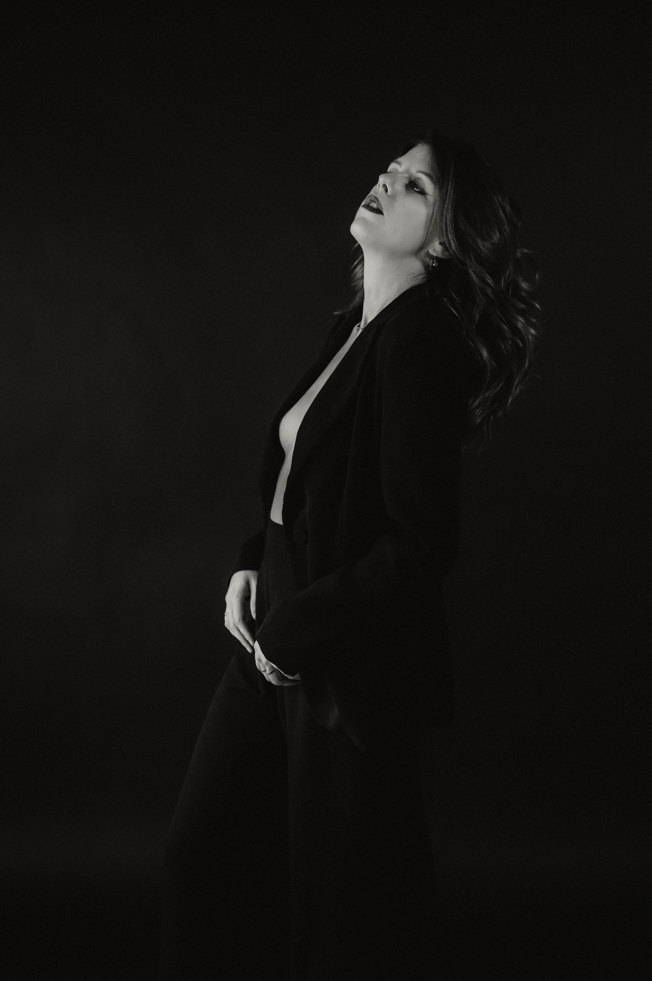 A black and white photo of a woman with dark wavy hair, wearing dark lipstick and a black jacket, posing against a dark background. She has her head tilted back slightly, with her lips parted and eyes looking upward.