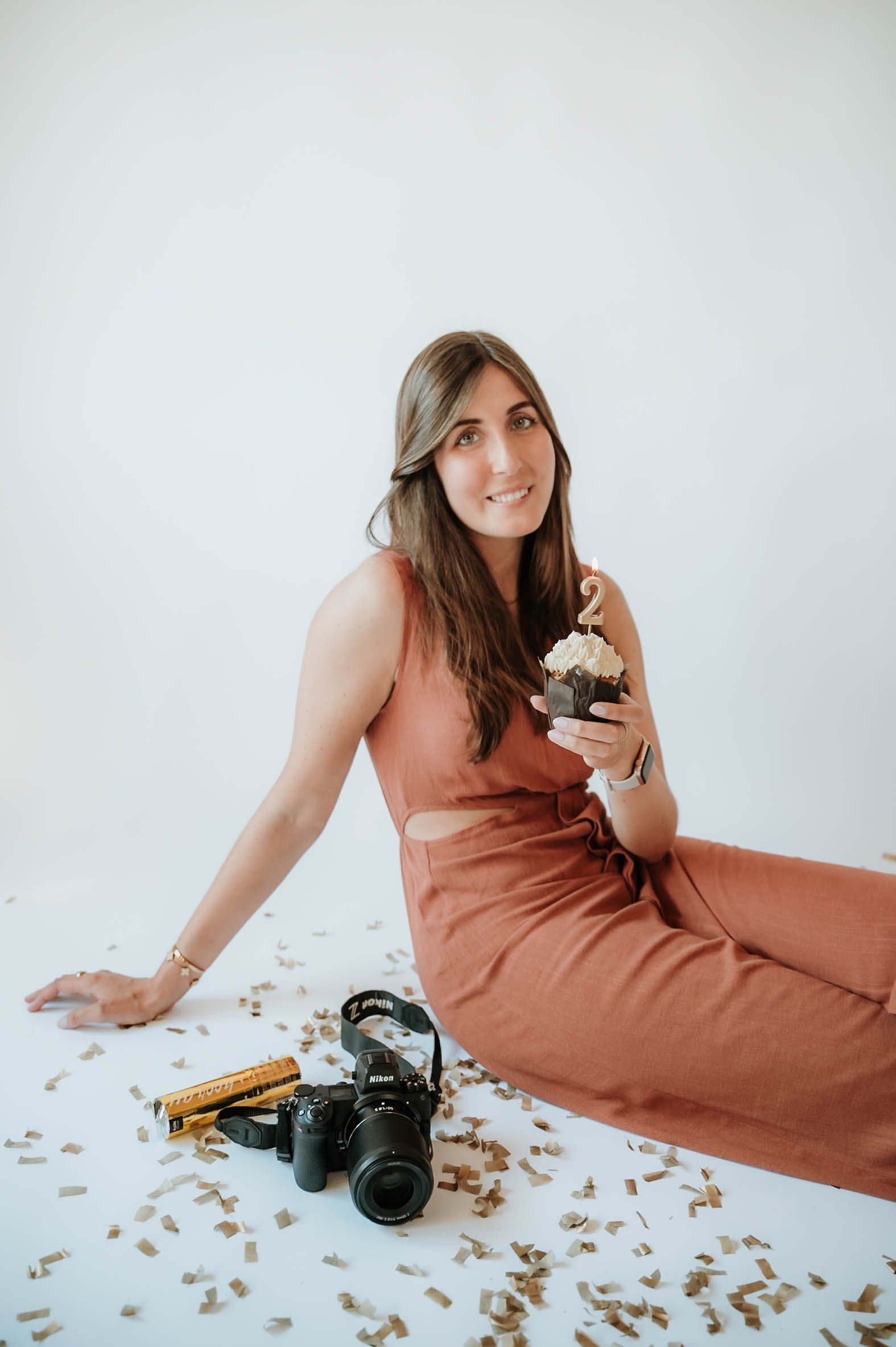 A woman with long brown hair sitting on a white floor surrounded by scattered beige confetti pieces. She is holding a cupcake with a candle shaped as the number 2. There is a Nikon camera, a box, and other items on the floor around her. She is wearin
