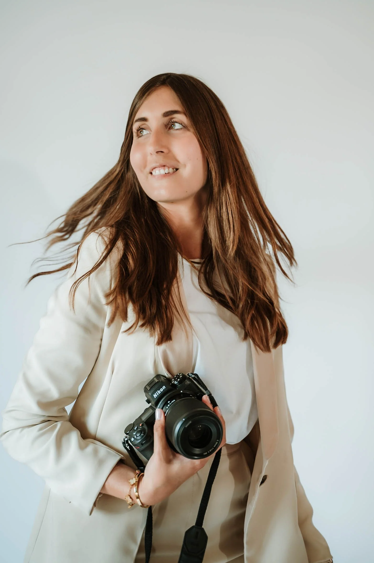 A woman with long brown hair wearing a beige blazer and holding a Nikon camera. She is slightly smiling and looking to her left, with her hair moving as if a breeze is blowing.