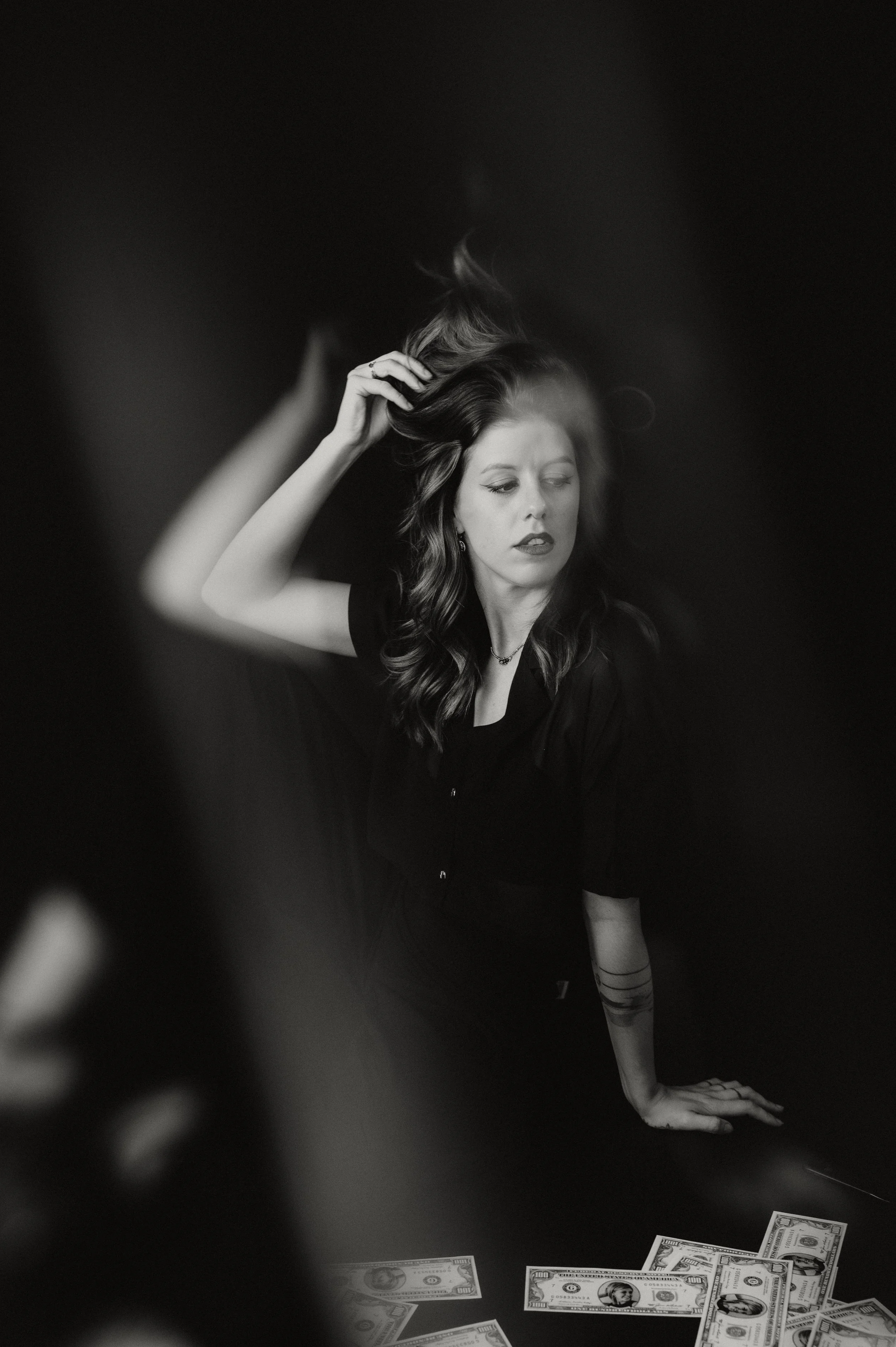 A woman with long wavy hair wearing a black top sitting on the floor, touching her hair, with scattered U.S. dollar bills on the floor in front of her, all in black and white.