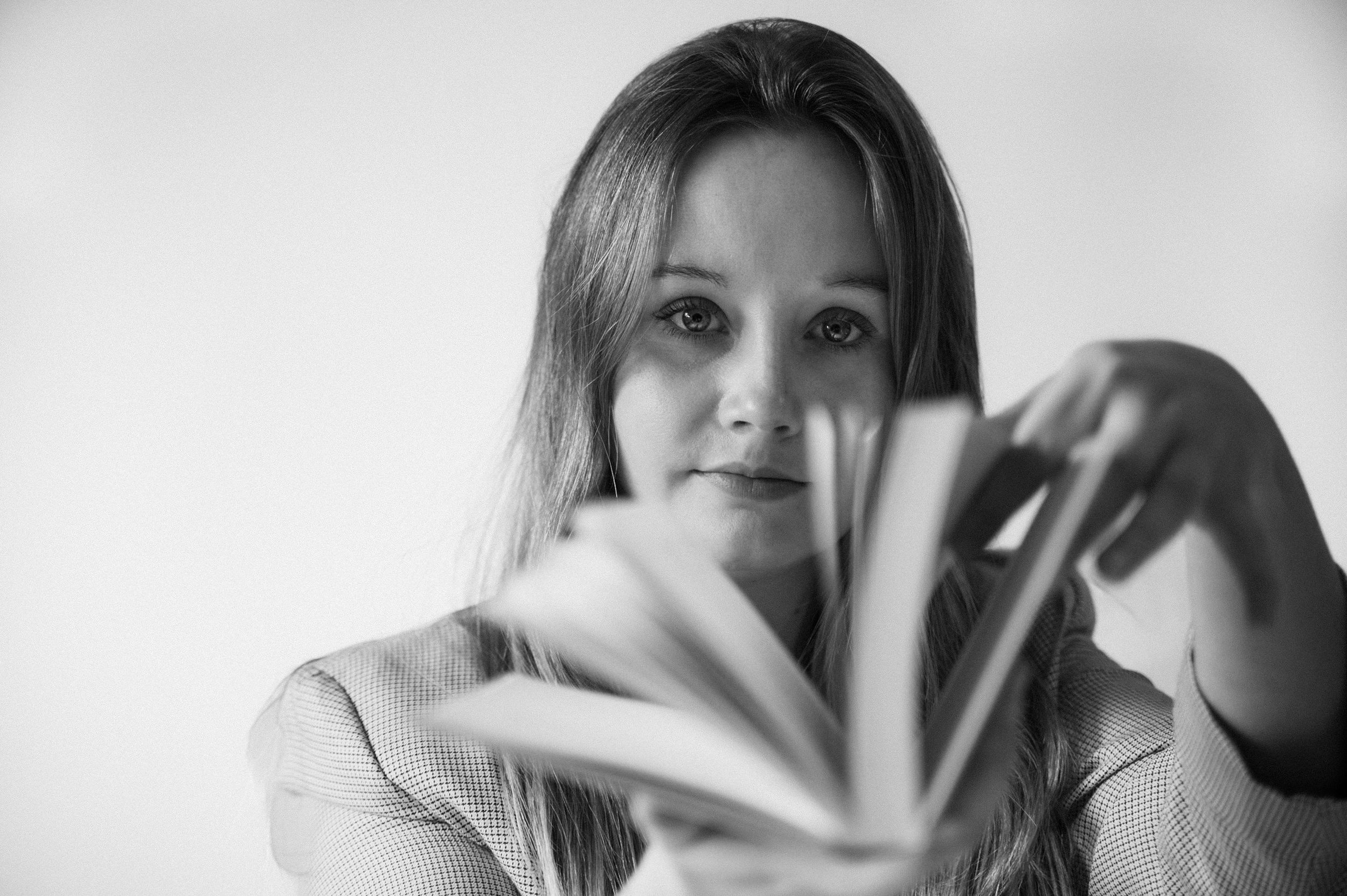 Black and white close-up of a woman with long hair, looking at the camera, and holding a book or magazine in front of her.