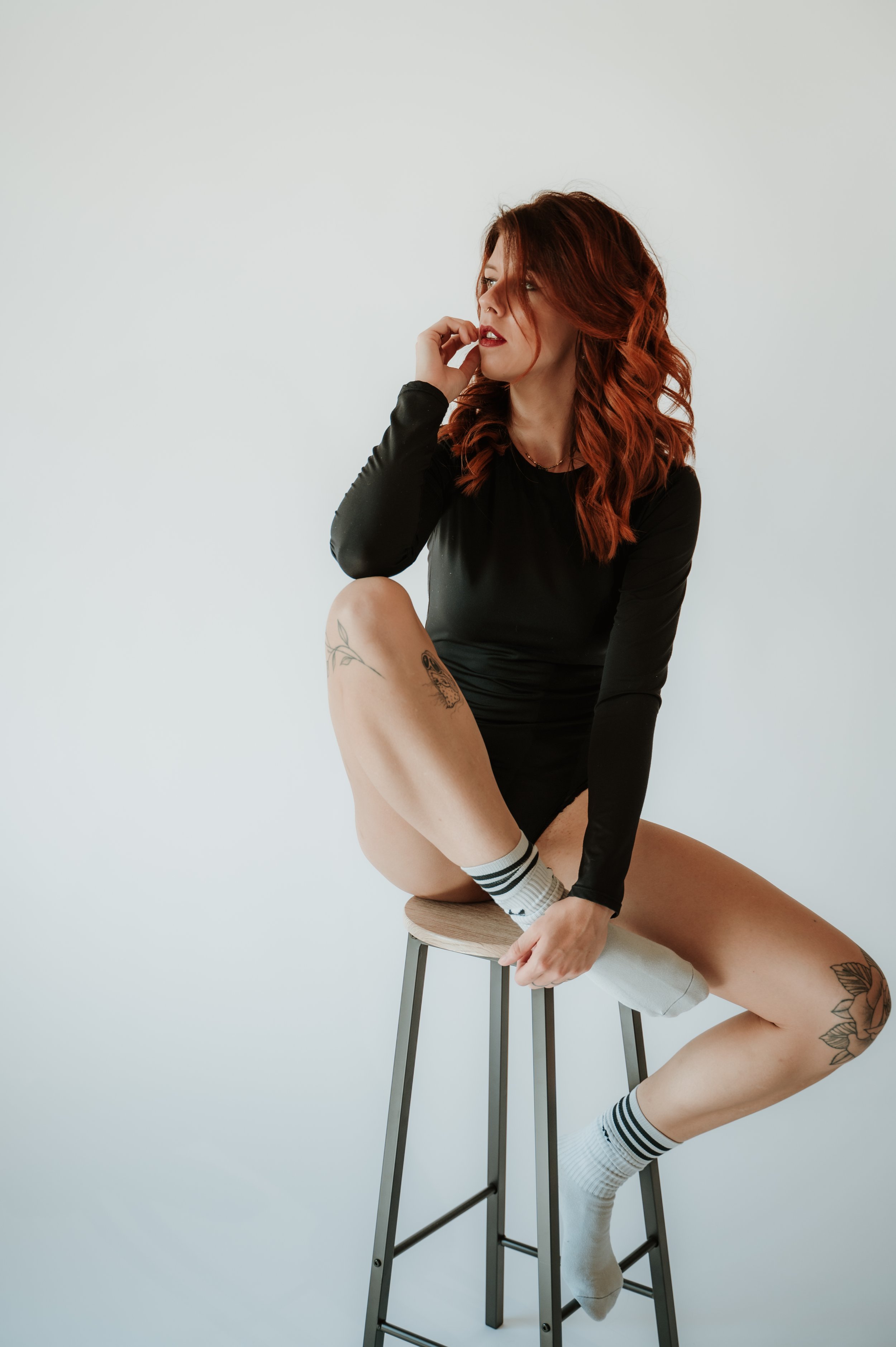 A woman with red hair sitting on a bar stool, wearing a black long-sleeve top, shorts, and striped socks, touching her face and looking to the left against a plain white background.