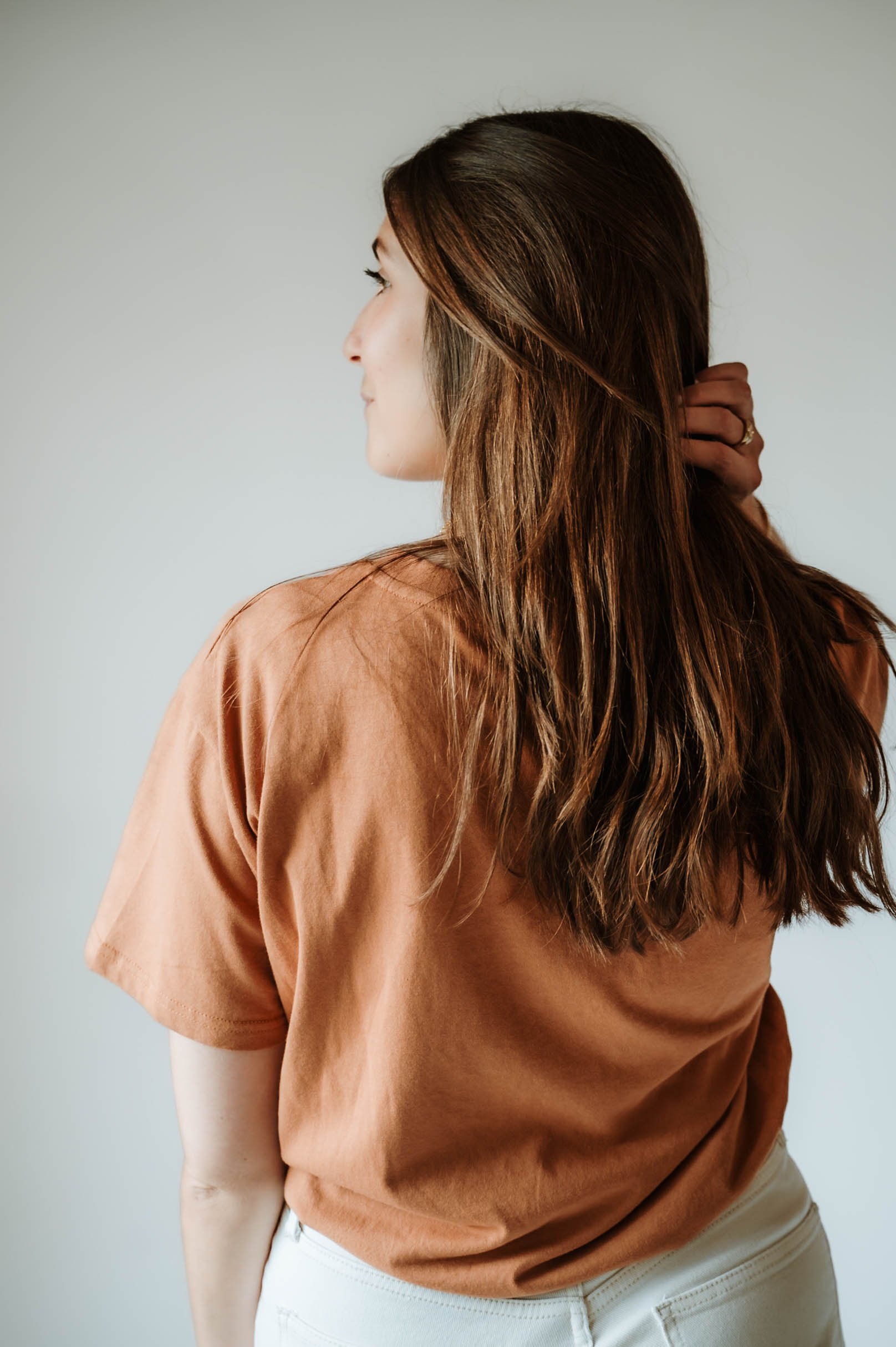Side view of a woman with long brown hair wearing a tan t-shirt and white pants, holding her hair behind her head, against a light gray background.