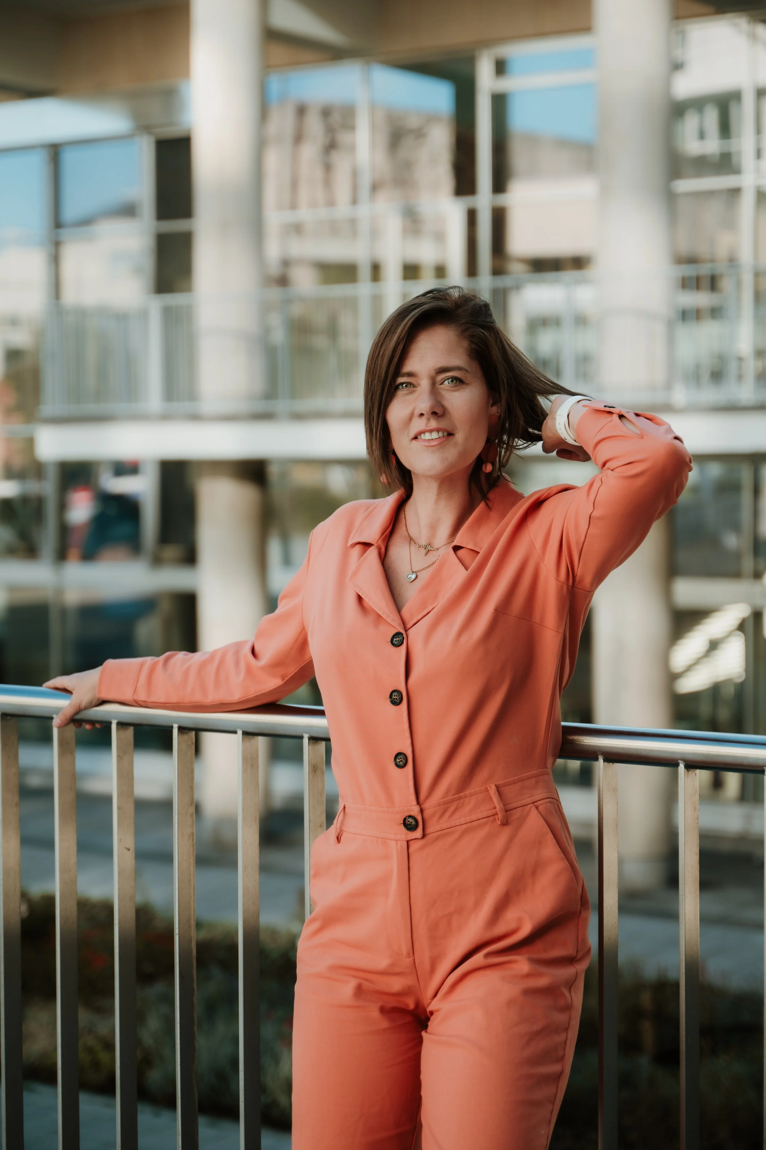 A woman with shoulder-length dark hair wearing a coral-colored jumpsuit, standing outdoors next to a metal railing, with a modern building featuring large glass windows and structural columns in the background.