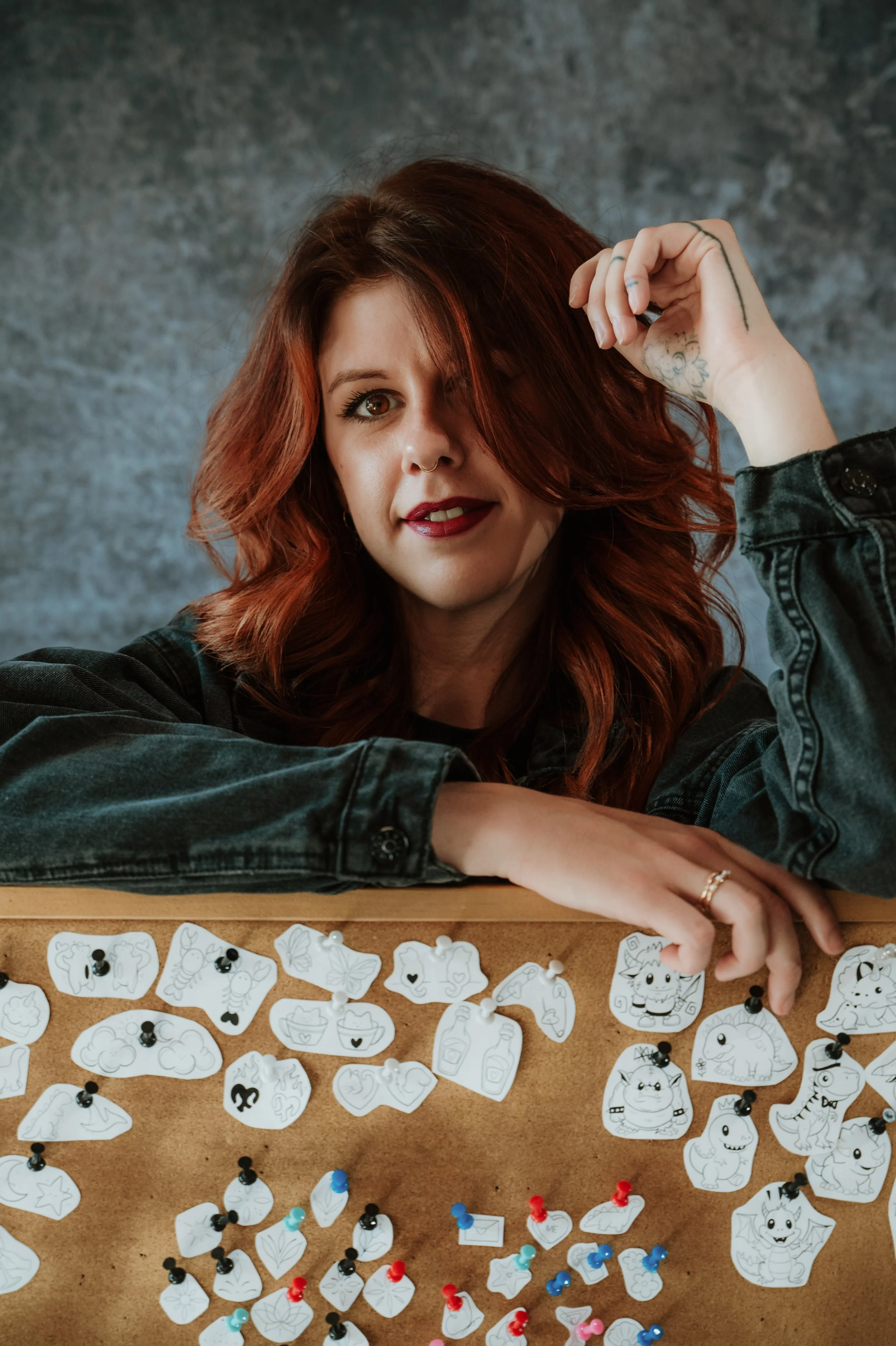 A woman with red hair and a nose ring sitting at a table with black and white cartoon-themed paper cutouts and colored push pins.