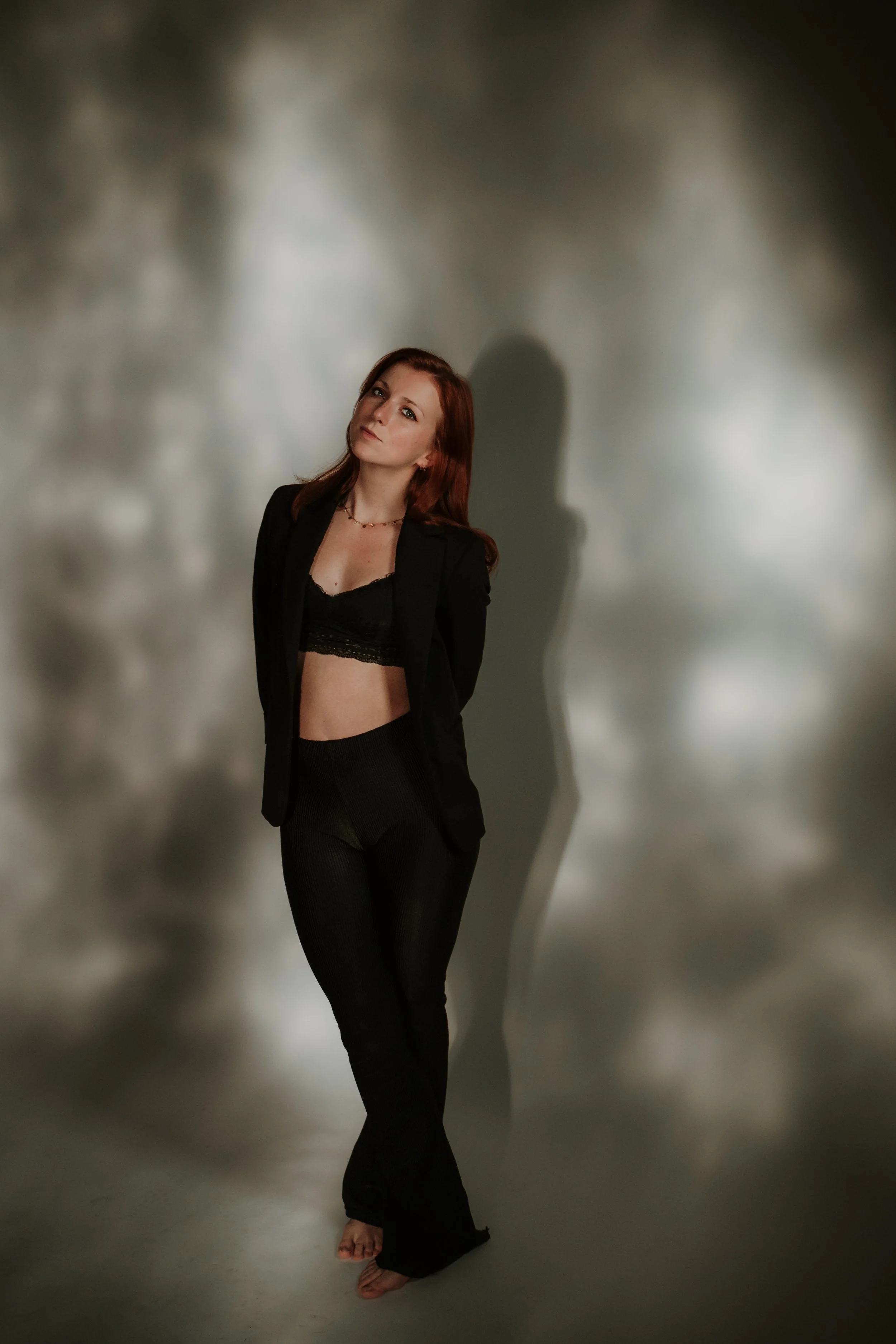 Woman with long reddish hair, wearing a black blazer and black lace crop top, standing against a blurry, cloudy background, with her shadow cast on the wall behind her.
