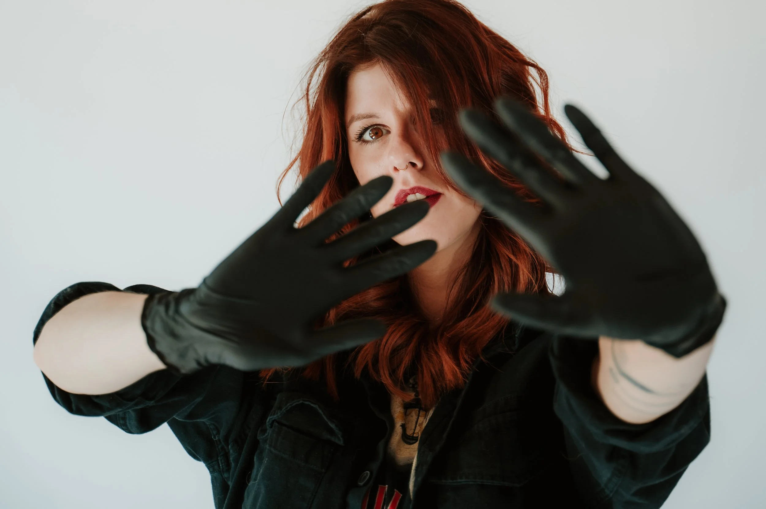 Young woman with red hair and a nose ring, wearing black gloves and a dark jacket, making a playful gesture towards the camera against a plain light-colored background.