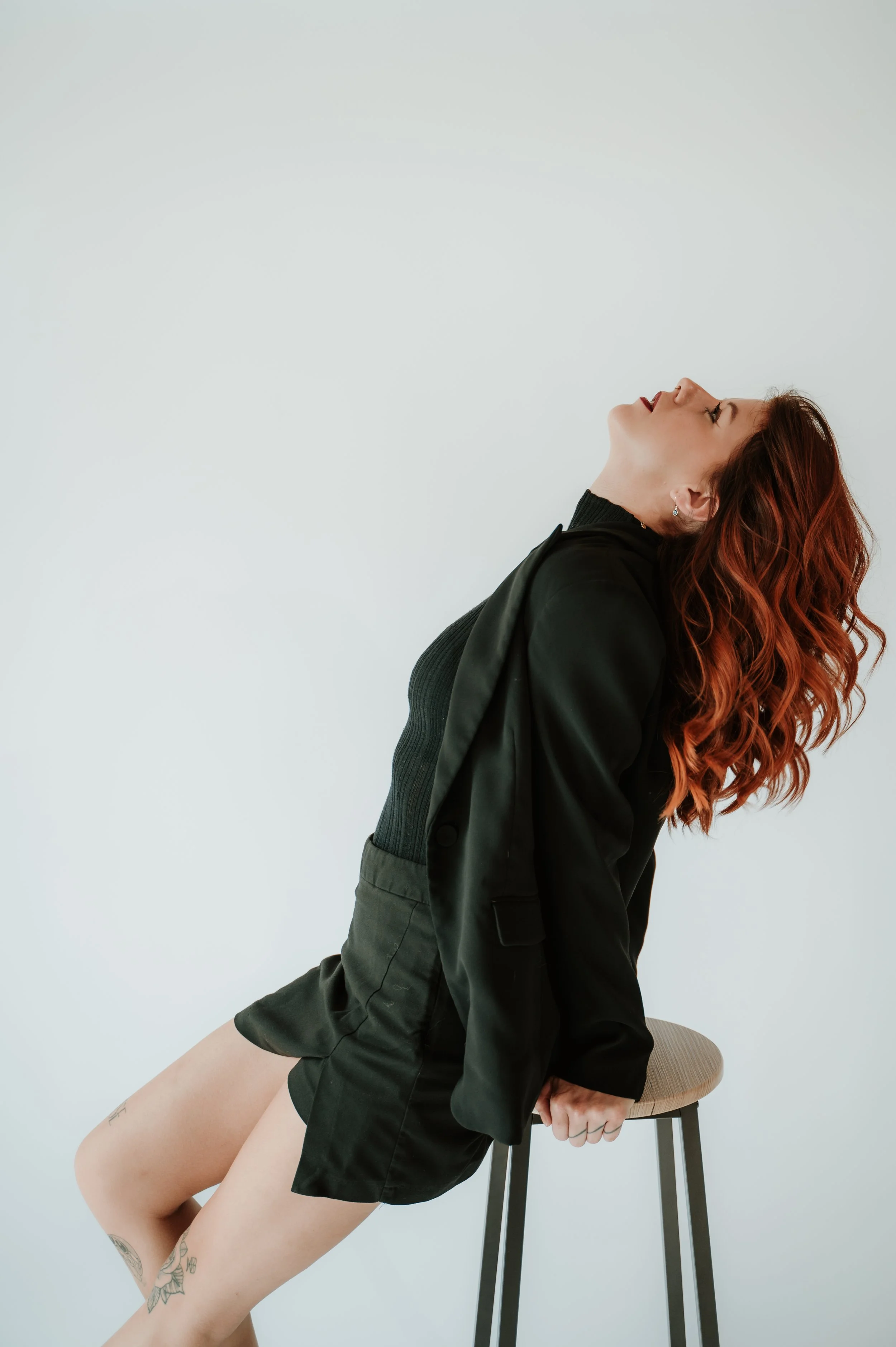A woman with red wavy hair, wearing a black turtleneck and blazer, is posed with her head tilted back and eyes closed, sitting on a stool. She appears to be engaged in a pose or movement, with her legs slightly apart and one hand resting on the stool