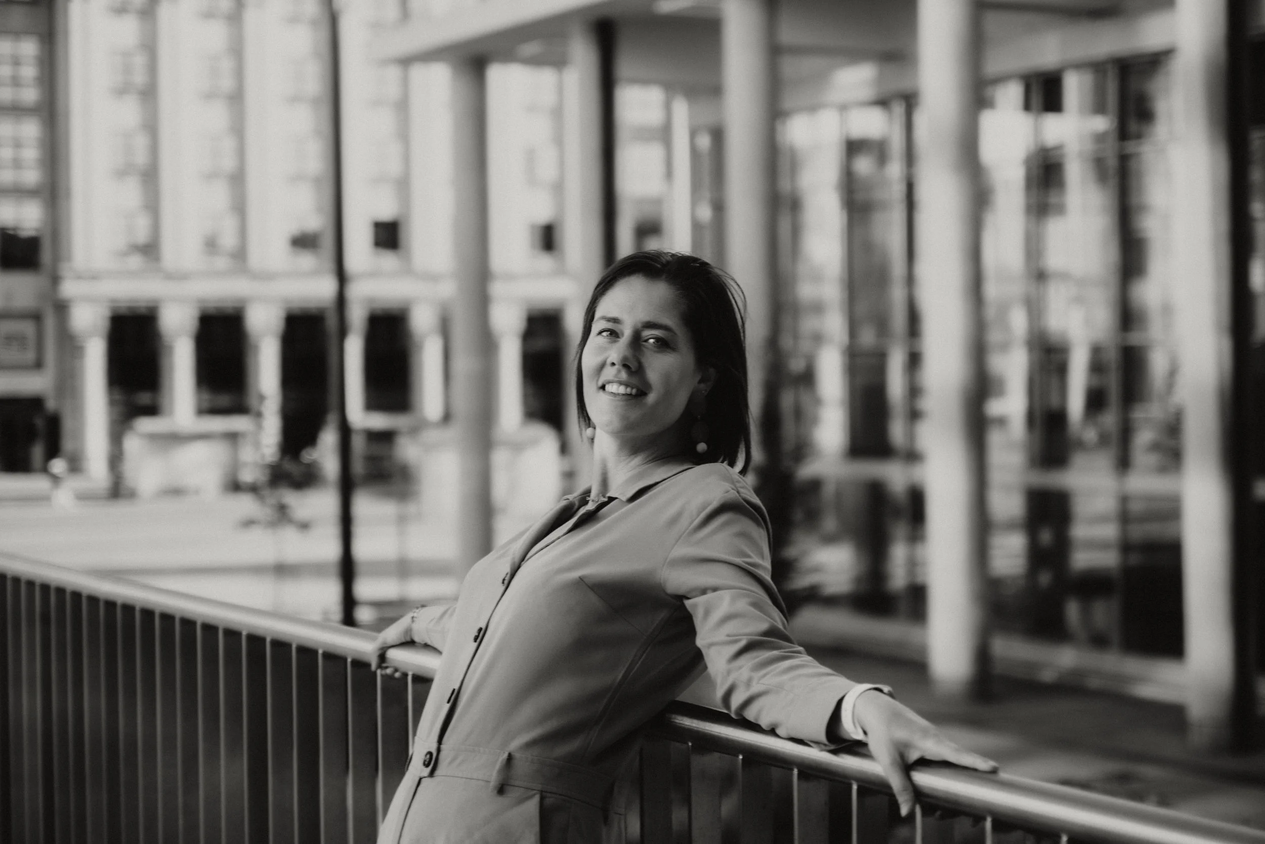 A woman standing outdoors near a railing, smiling, with a background of modern multi-story buildings.