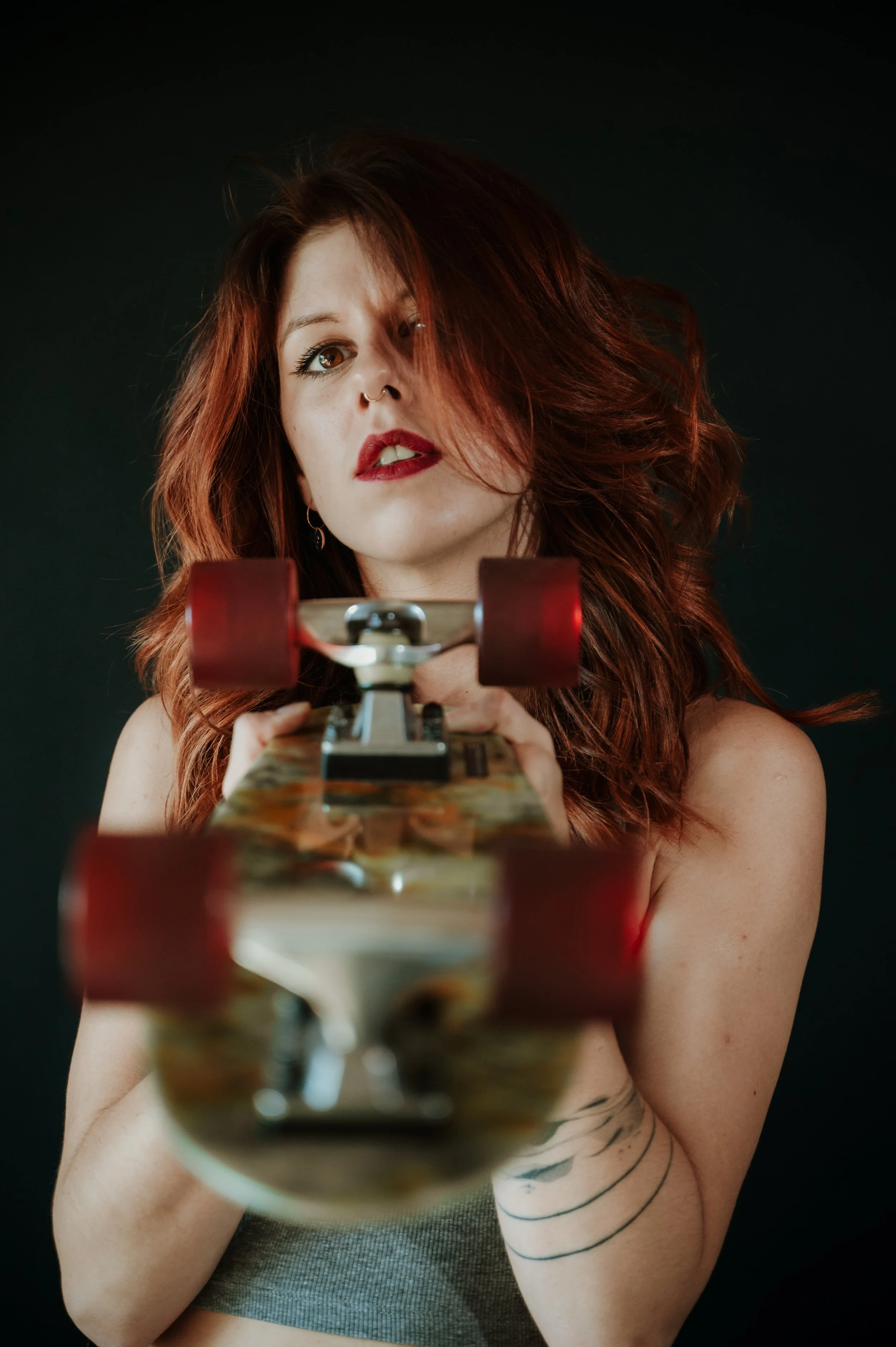 Young woman with red hair and tattoos on her arm, holding a skateboard and looking into the camera against a black background.