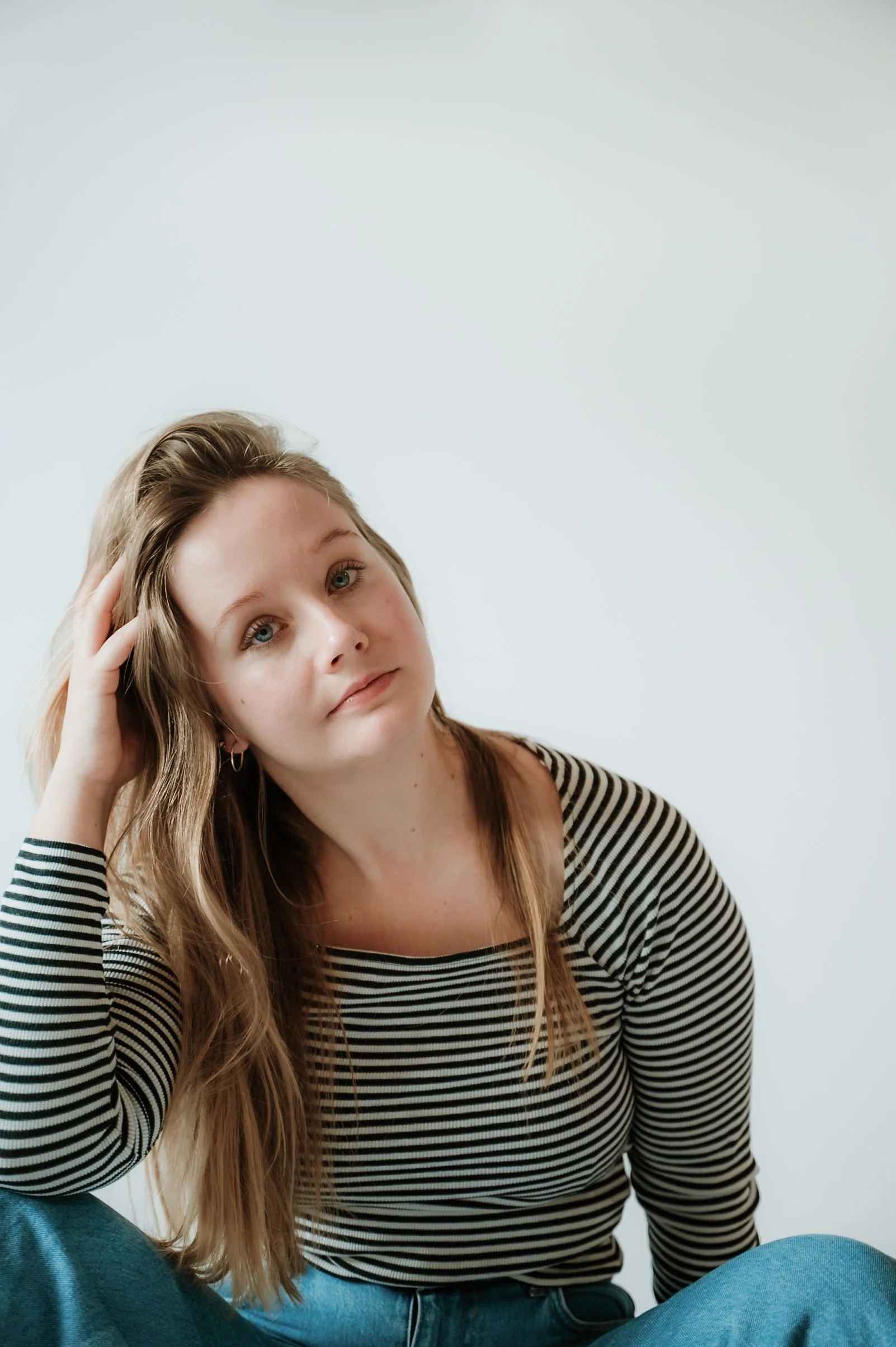 A young woman with long, wavy light brown hair, blue eyes, and fair skin is sitting in front of a plain white wall. She is wearing a black and white striped shirt and blue jeans. She is resting her right hand on her head and has a contemplative expre