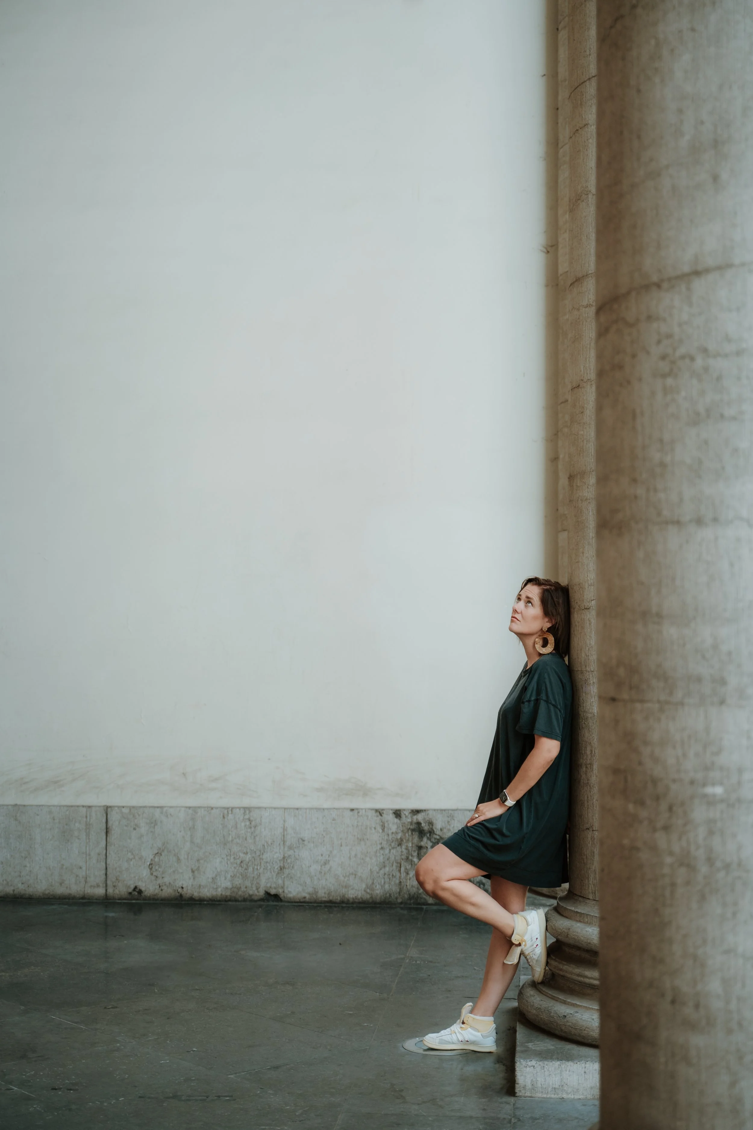 Woman in green dress leaning against a stone column in an empty room.