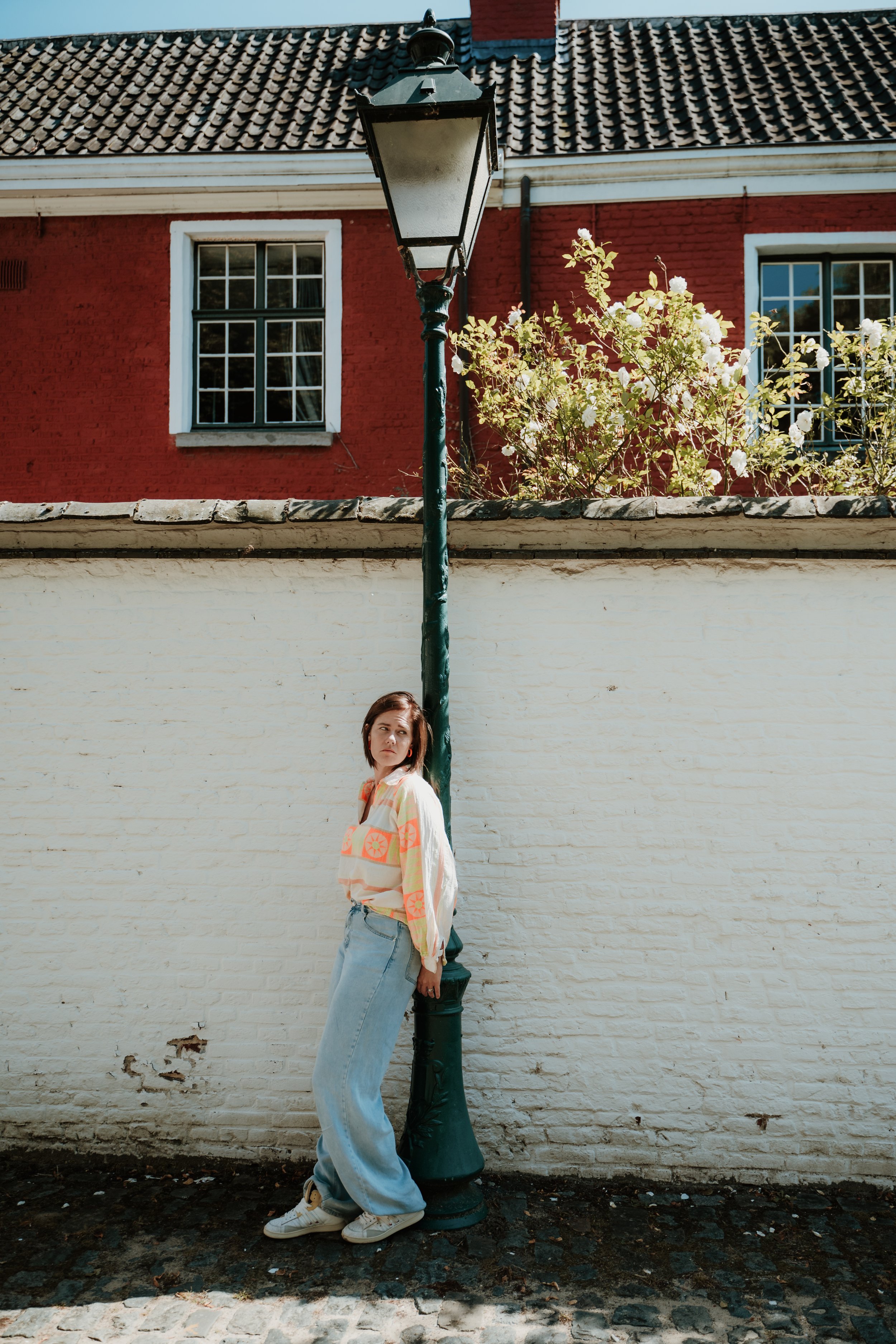 A woman with short hair and light skin leaning against a black streetlamp in front of a white brick wall. She wears a colorful patterned shirt, light blue jeans, and white sneakers. Behind the woman is a red brick house with white window frames and a