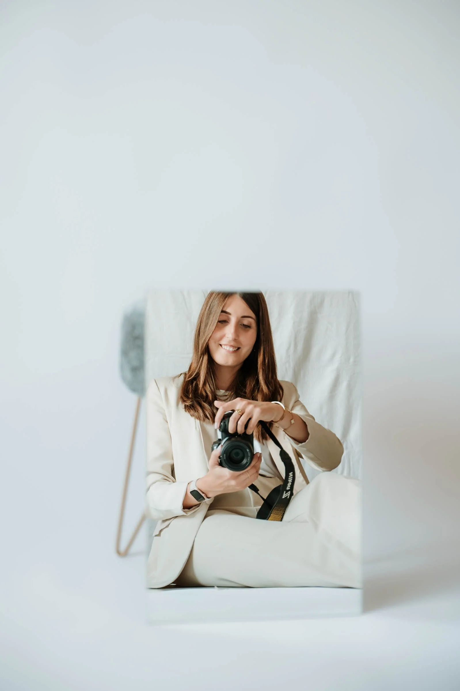 Woman with long brown hair taking a selfie with a DSLR camera while smiling, seen through a mirror, wearing a beige blazer, sitting on a bed with white linens in a minimal room.