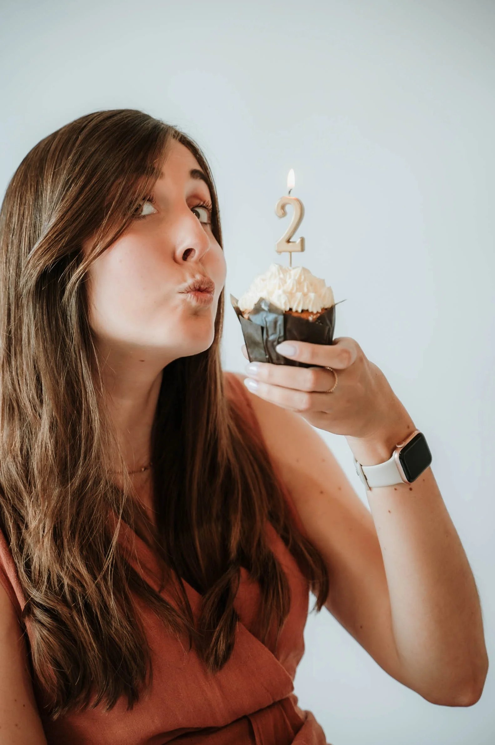 A woman with long brown hair wearing a brown dress and a white smartwatch holding a cupcake with white frosting and a birthday candle shaped like the number 2, blowing out the candle.