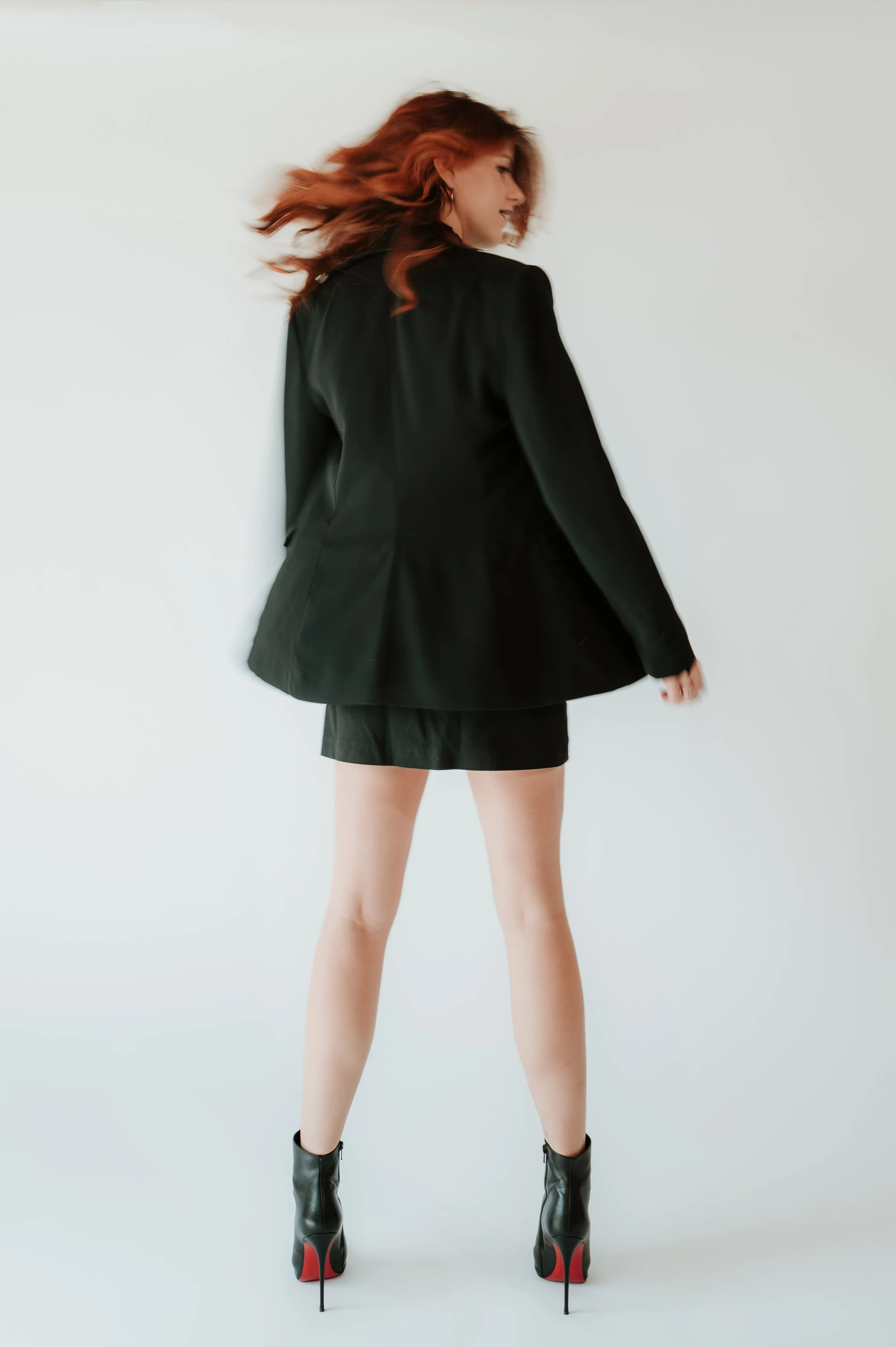 A woman with red hair wearing a black blazer, black mini skirt, and black high-heeled boots with red soles, standing against a white background.