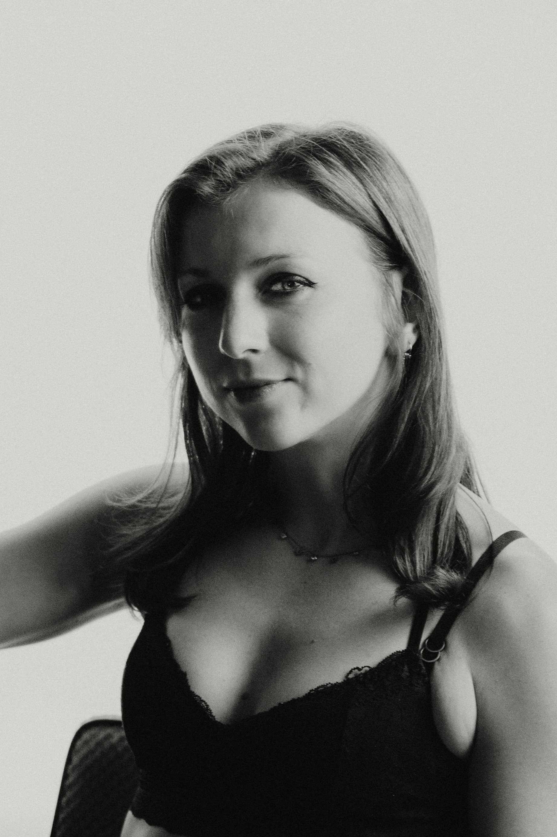 Black and white photo of a woman taking a selfie, wearing a lacy black top and a necklace, with long hair styled in loose waves, looking at the camera with a slight smile.