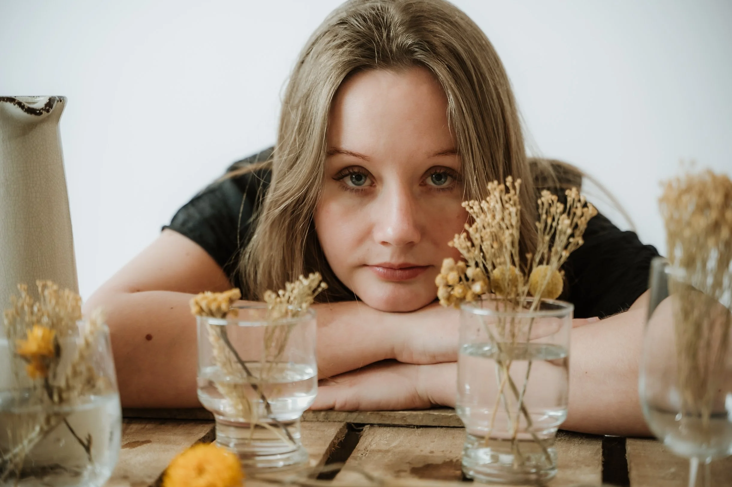 A young woman with light brown hair and blue eyes resting her chin on her arms, looking directly at the camera, surrounded by small glass cups filled with flowers or dried plants on a wooden table.