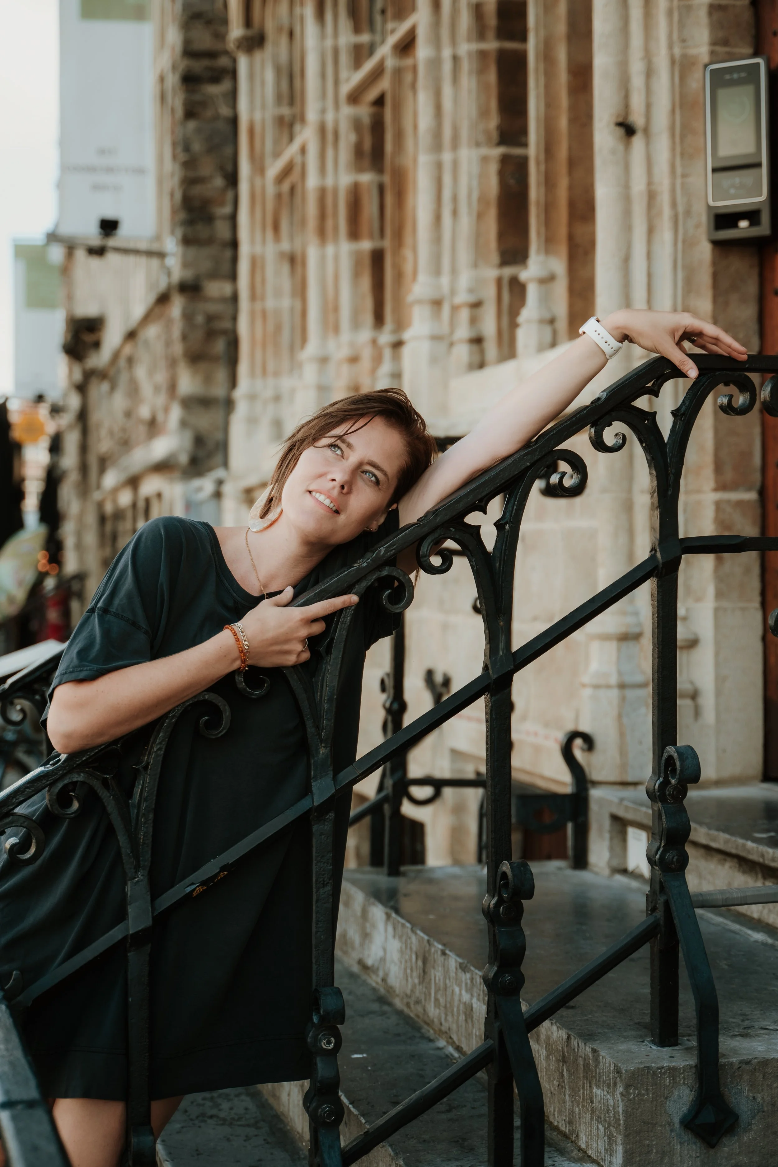 A woman leaning on an ornate black metal railing outside a building with historic architecture, wearing a black top and a white watch.