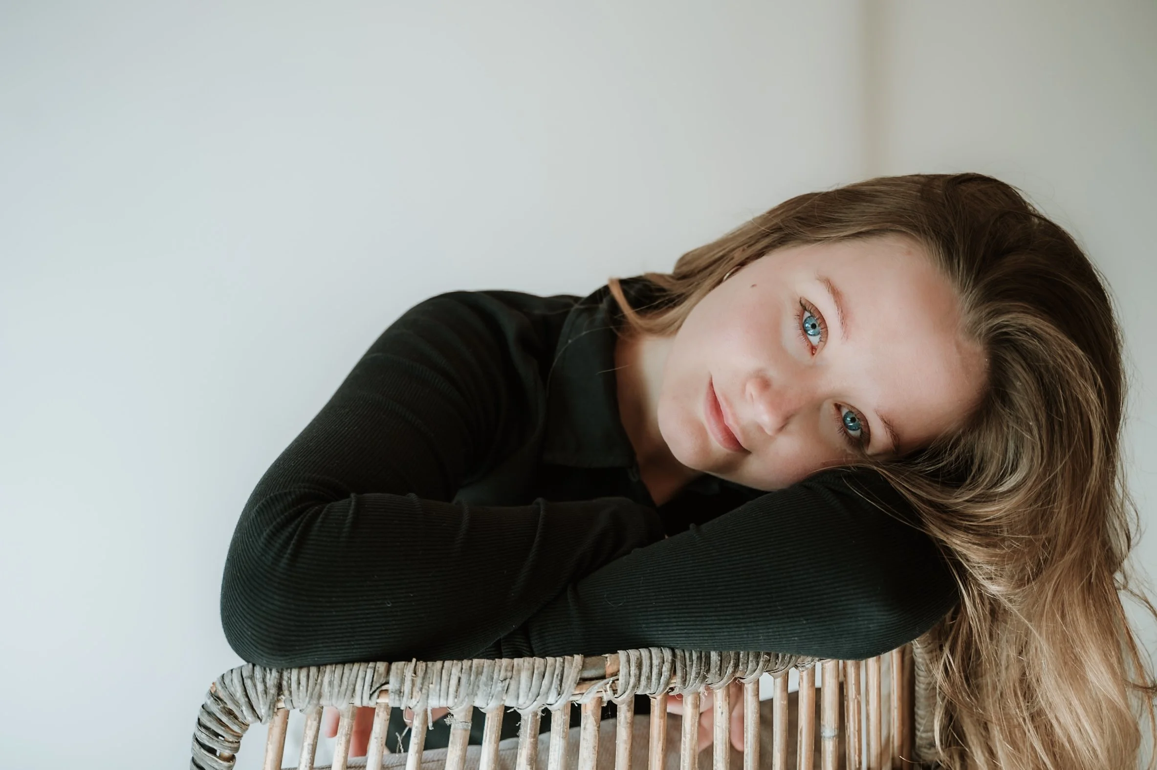 A young woman with long, wavy brown hair and blue eyes, smiling softly, resting her head on her arms, which are folded on a wicker surface, against a plain white background.