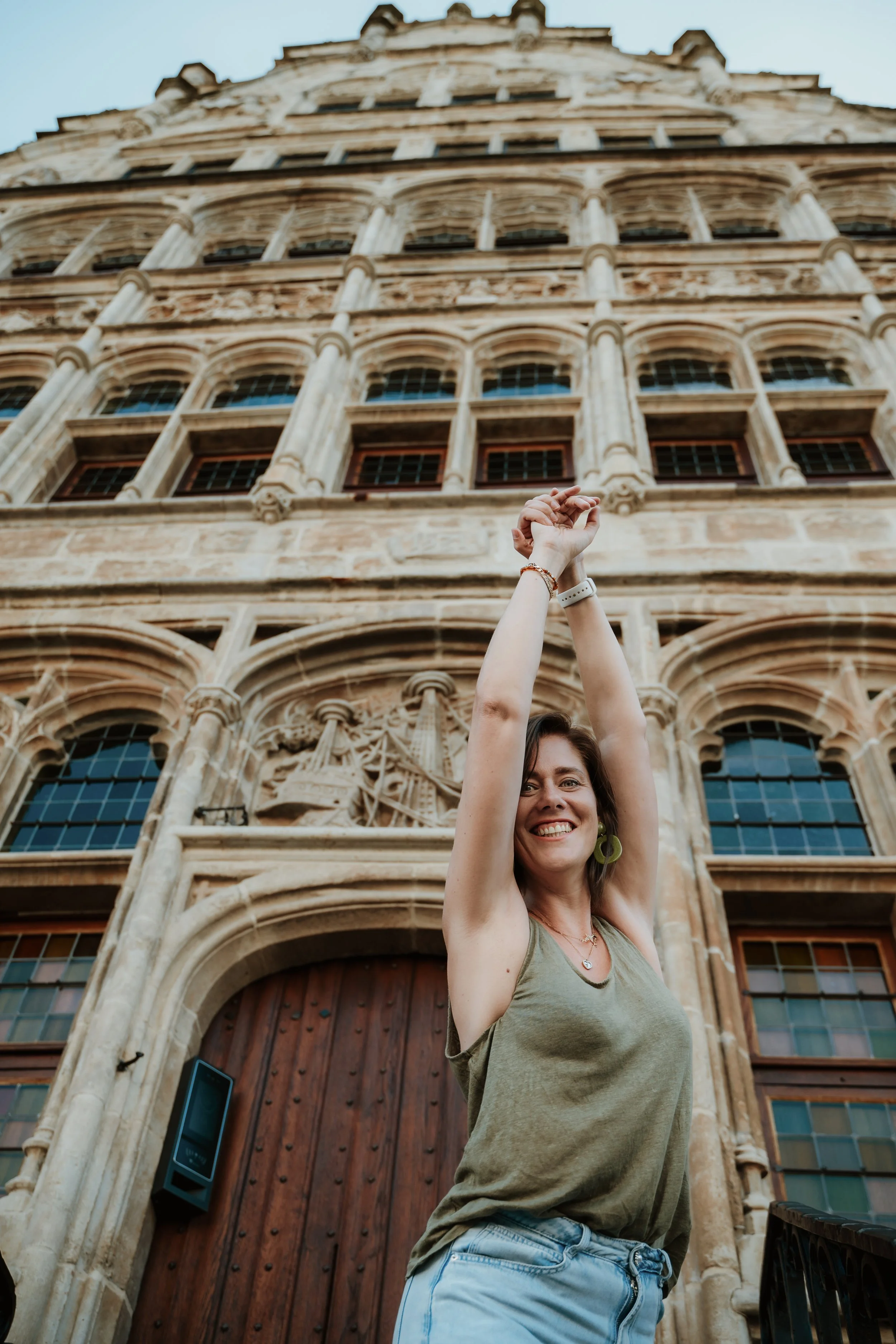 A woman smiling and raising her hands above her head in front of a historic, ornately decorated building with arched windows and carved stone details.