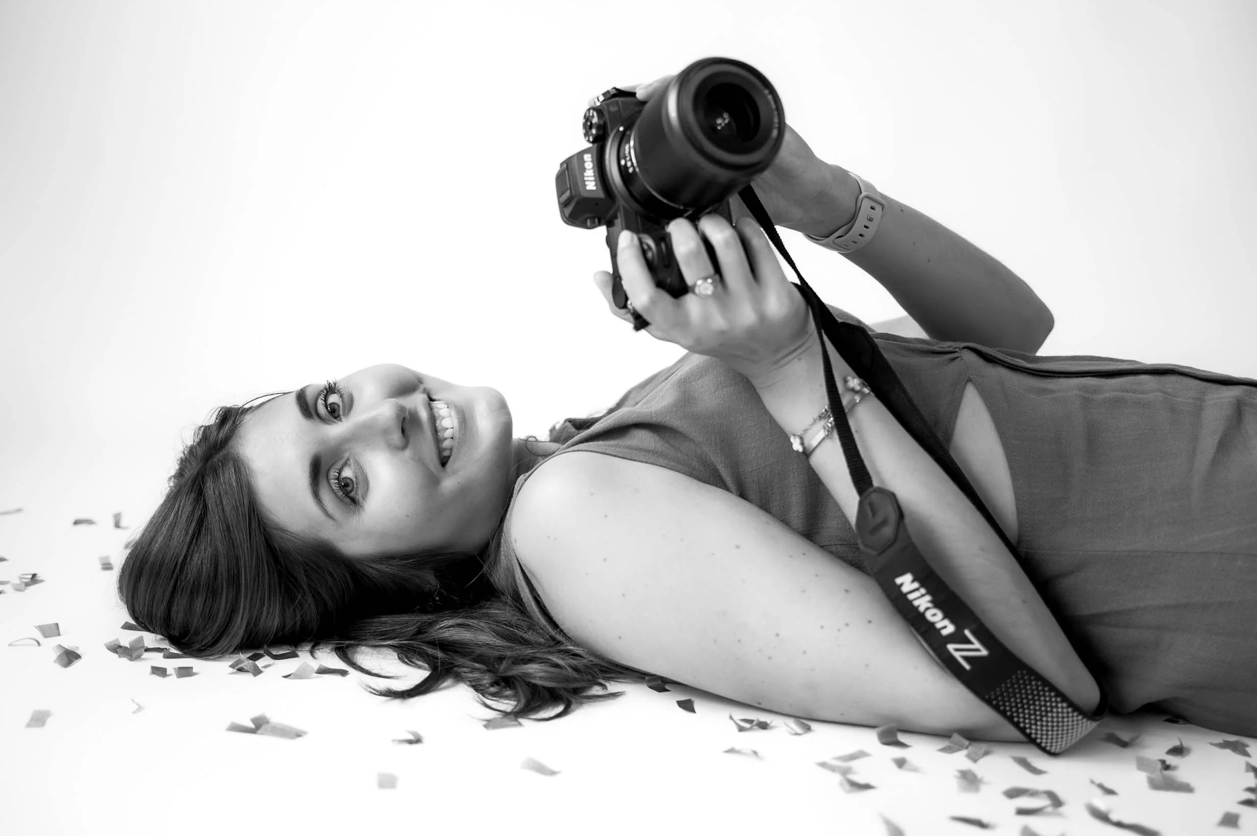 A woman lying on her side with her hair spread out, smiling, holding a Nikon DSLR camera, surrounded by scattered confetti on a plain backdrop.