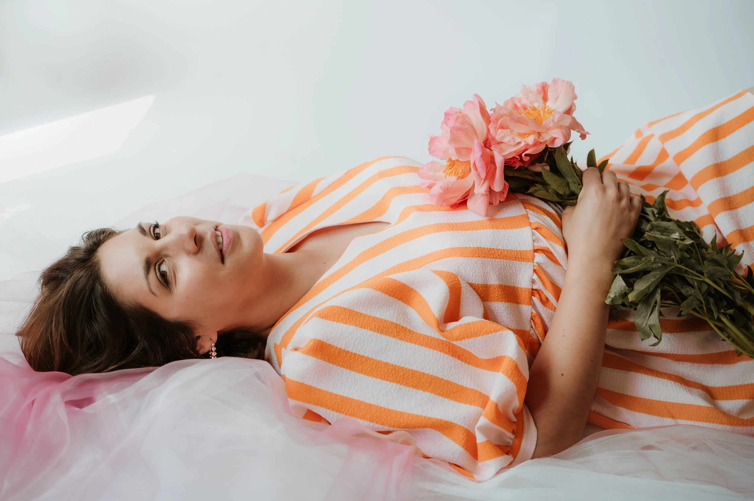 A woman lying on a bed holding a bouquet of pink peonies, wearing an orange and white striped dress, with pink tulle fabric on the bed.